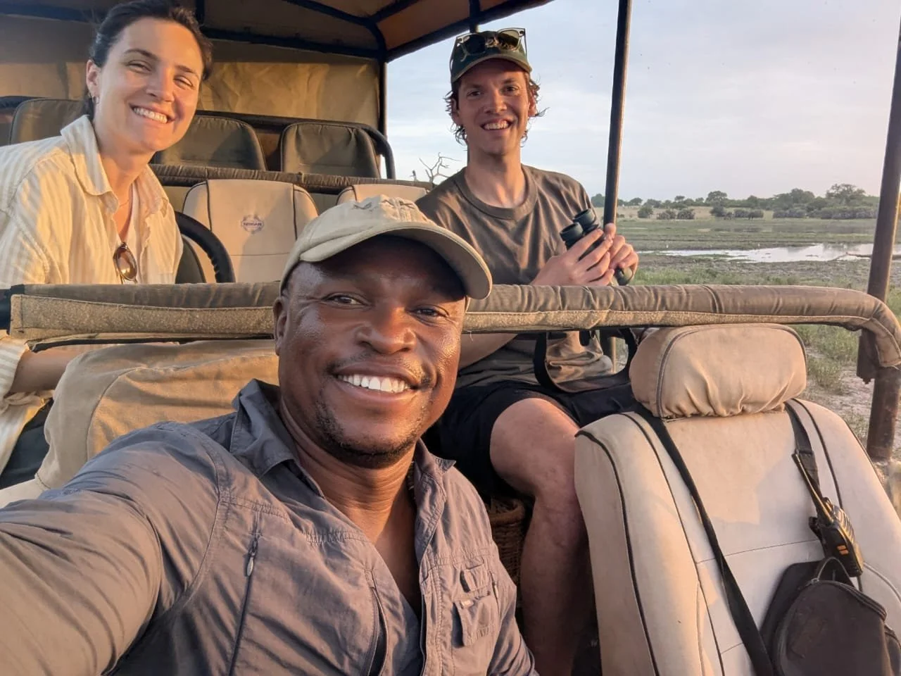 A group of four people on a safari vehicle taking a selfie in a savannah landscape with trees and water in the background at sunset.