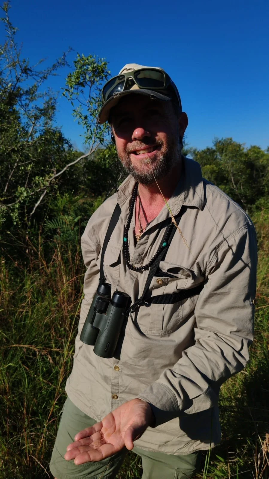 A smiling man in outdoor wildlife or nature attire, holding a small insect on his hand, with binoculars around his neck, in a green, bushy environment under a clear blue sky.