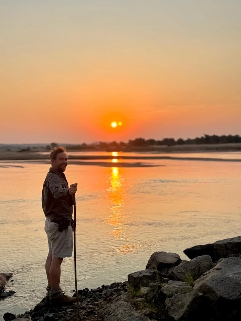 A man standing on rocks near a river during sunset, holding a walking stick and smiling at the camera.