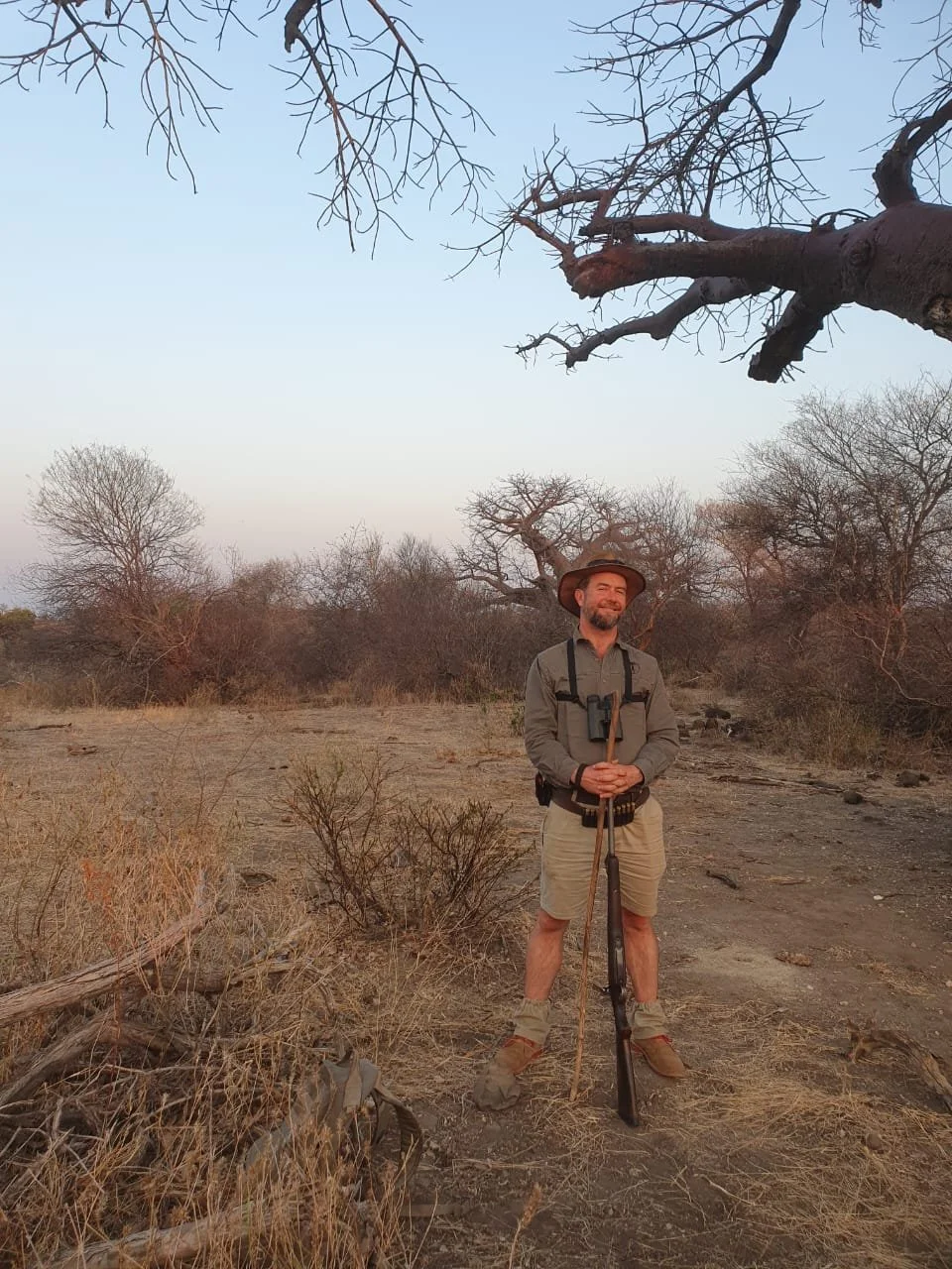 A man in safari attire standing outdoors in a dry, sparse landscape with leafless trees, holding a walking stick, smiling, with binoculars around his neck, under a large, leafless tree.
