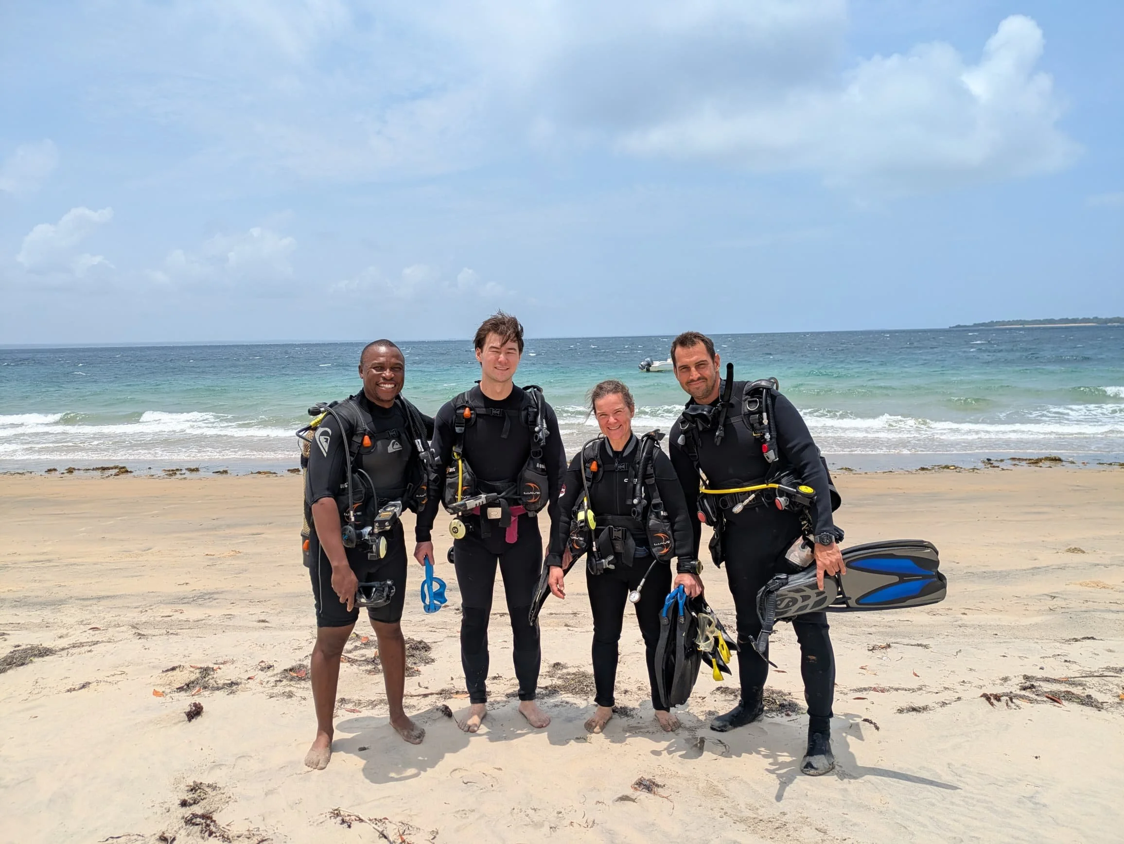 Four scuba divers wearing wetsuits and carrying scuba gear standing on a sandy beach with the ocean and a cloudy sky in the background.