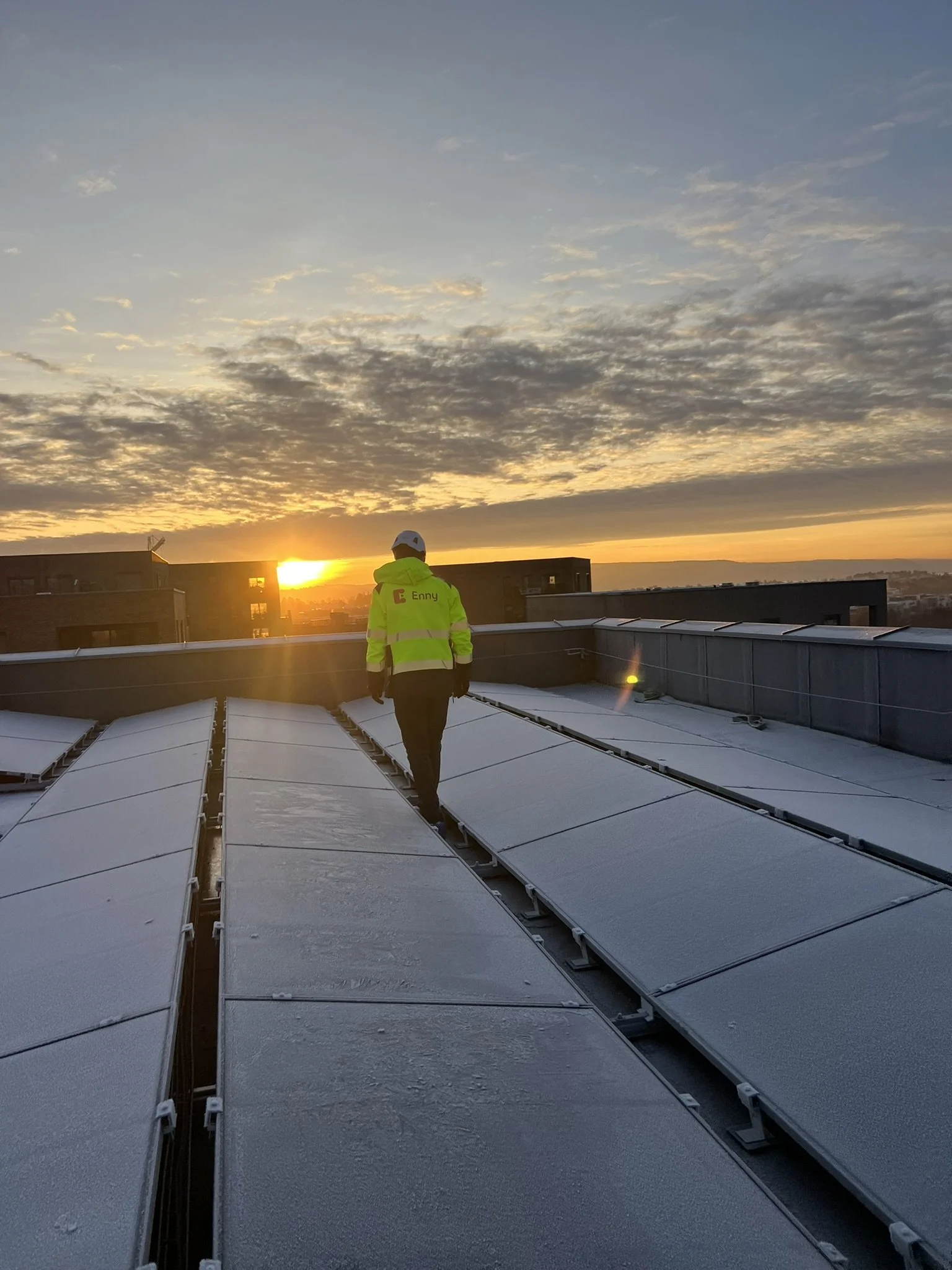 En person i en gul vest med logo står på taket av en bygning, med solnedgang og skyer i bakgrunnen.