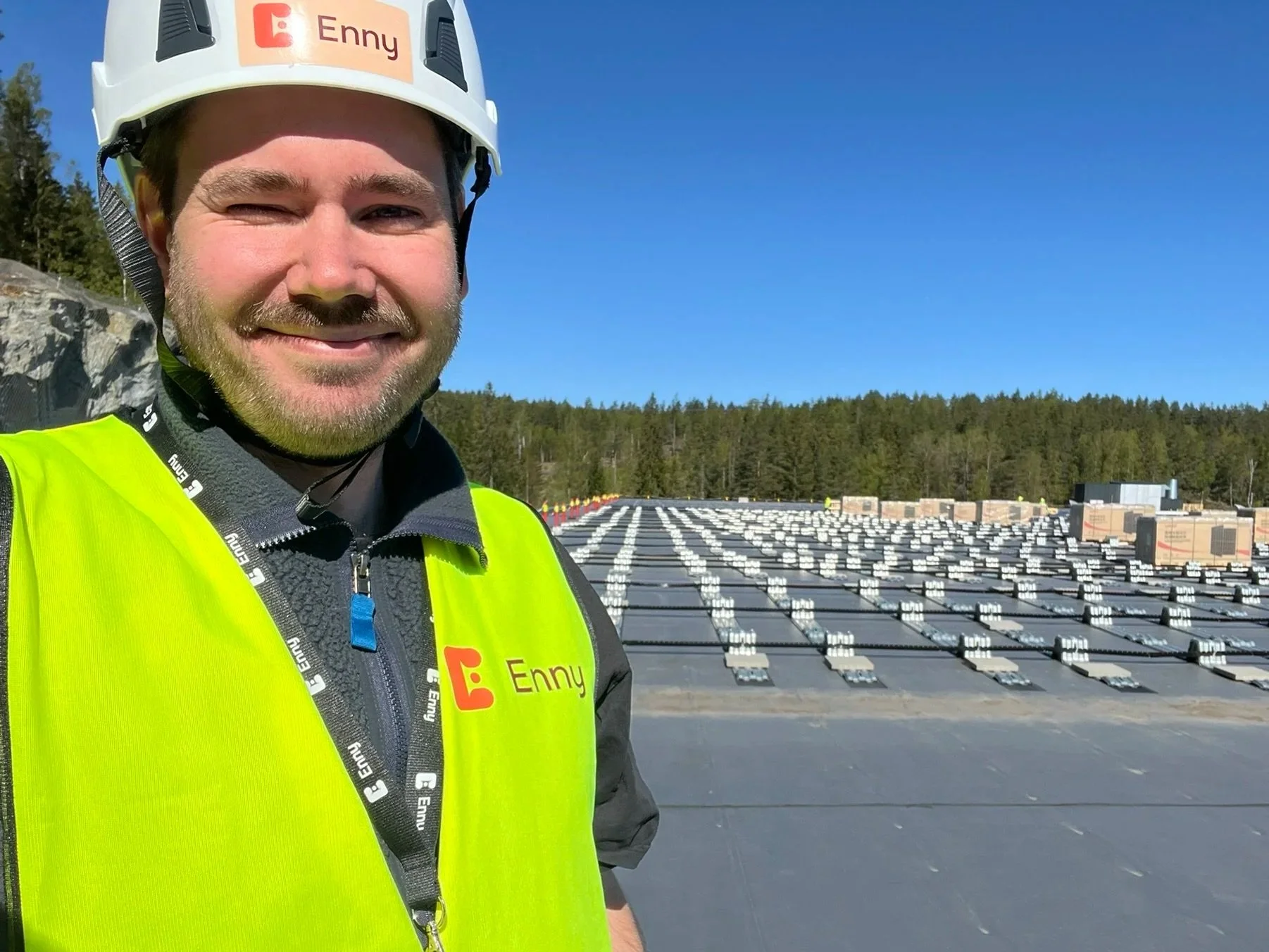 En mann som ser glad ut, iført hjelm og høy synlig vest, står foran en rekke med solcellepaneler på taket av en bygning, med skog og klar blå himmel i bakgrunnen.