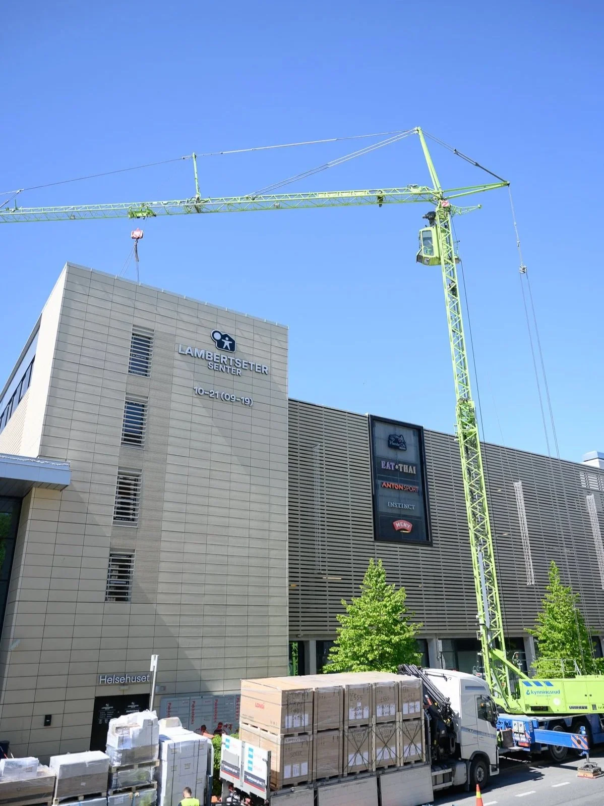 Bild av Lambertseter Sentrum, en gaffeltruck som lyfter byggmaterial och lastbilar parkerade framför en modern byggnad med skyltar och gröna träd.