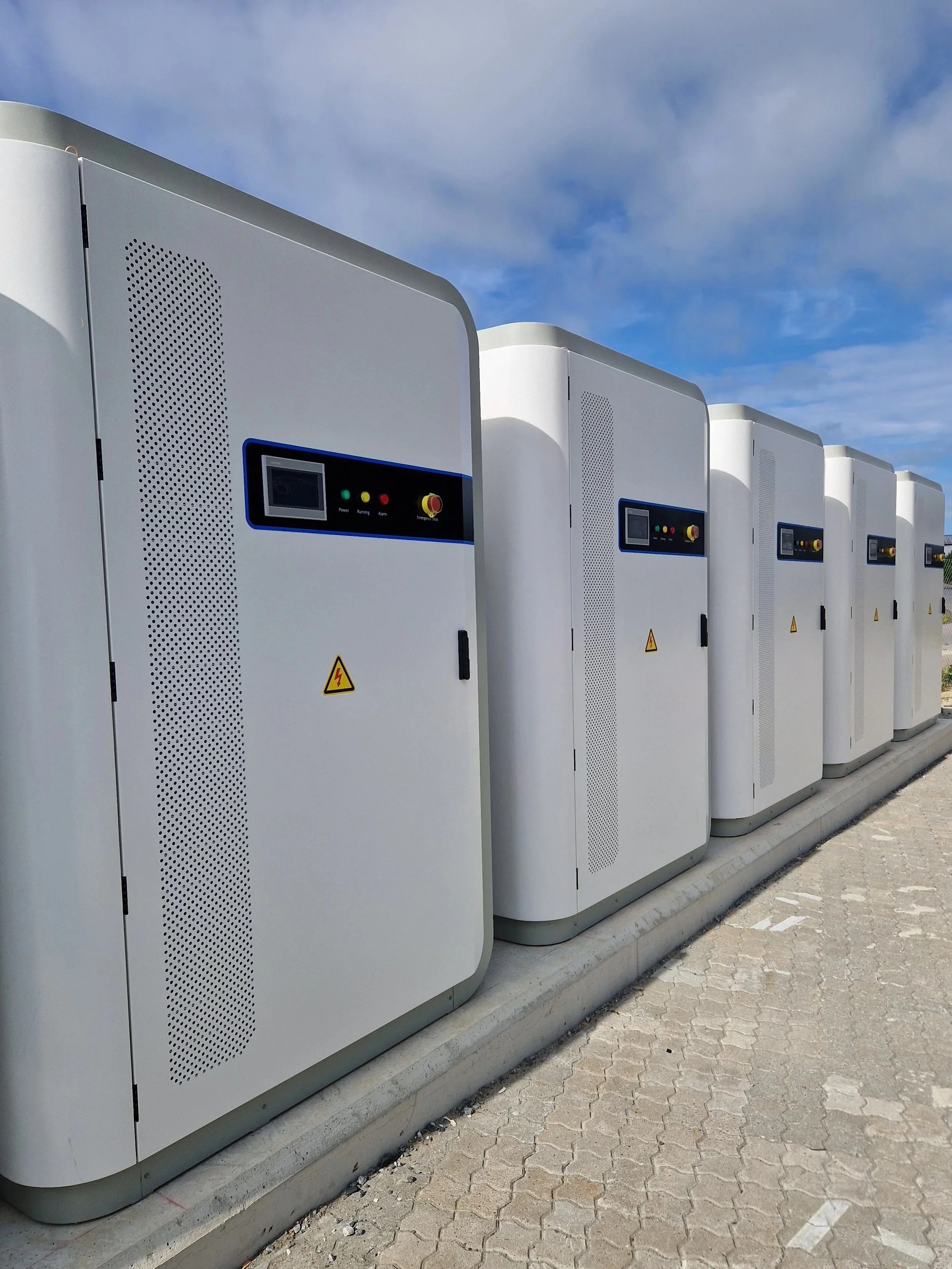 Fler elektriske ladebokser for elbiler plassert på en fortau, med en blå himmel og spredte skyer i bakgrunnen.