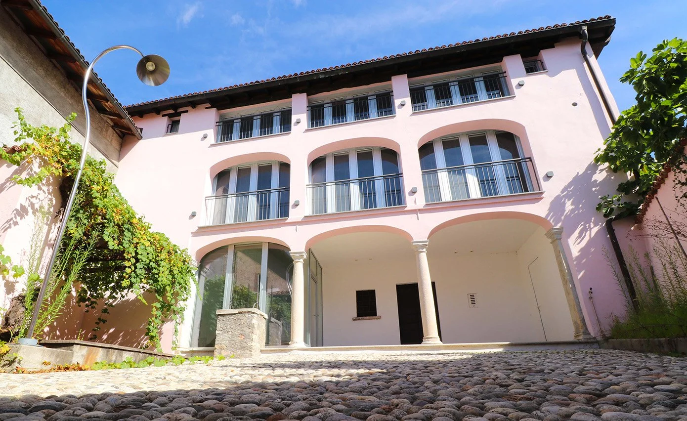 Front view of a multi-story pink house with arched windows and balconies, cobblestone walkway, and a curved street lamp on the left.