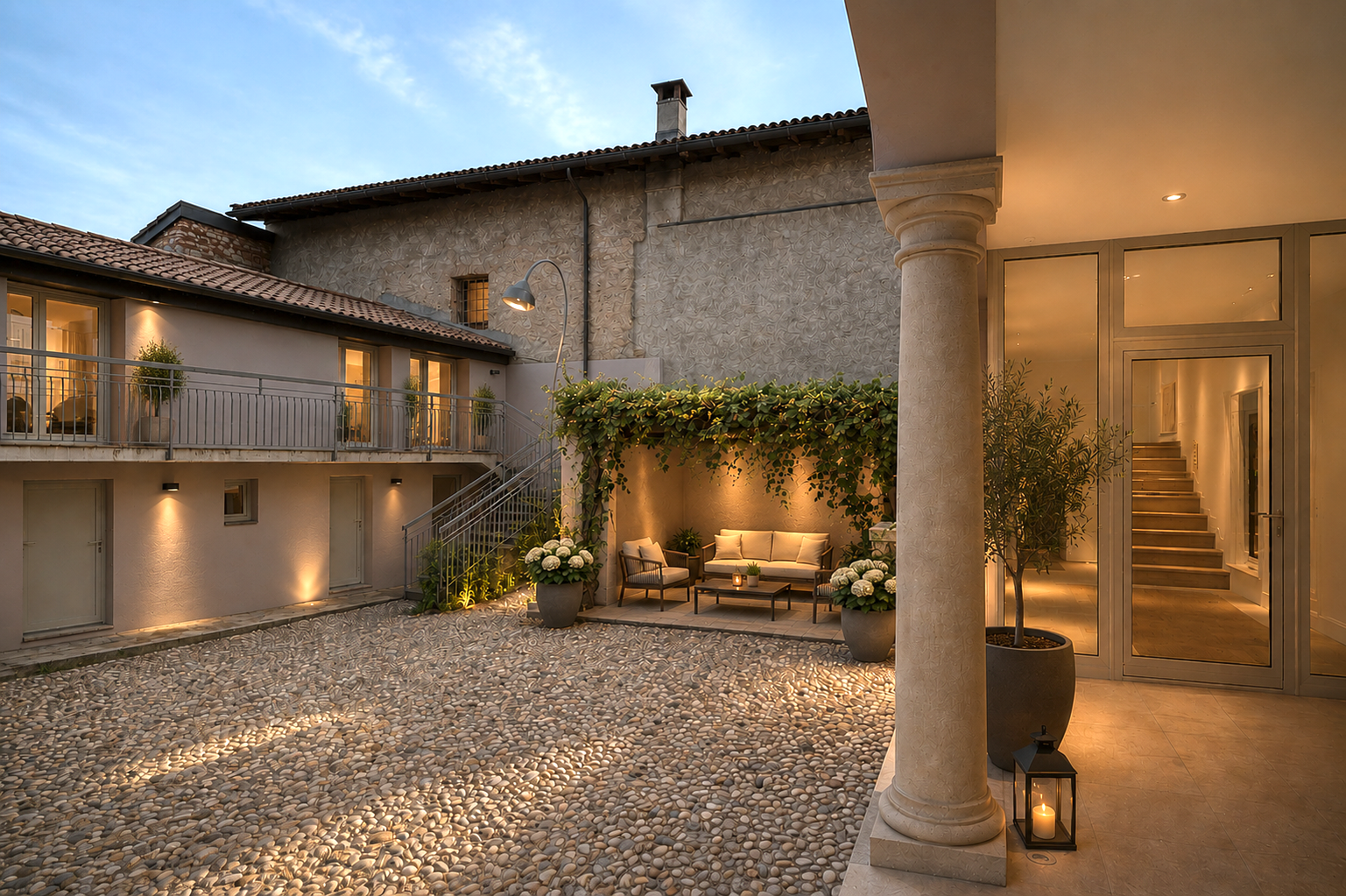 A cozy outdoor patio area at dusk with a couch, chair, and small table, decorated with potted plants, flowers, and candles, surrounded by a building with stone textures, windows with warm lighting, and a stone-tiled courtyard.