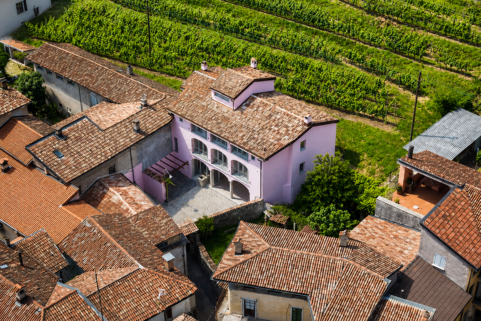 Aerial view of a neighborhood with terracotta-tiled roofs and a pink building, adjacent to a lush green vineyard.