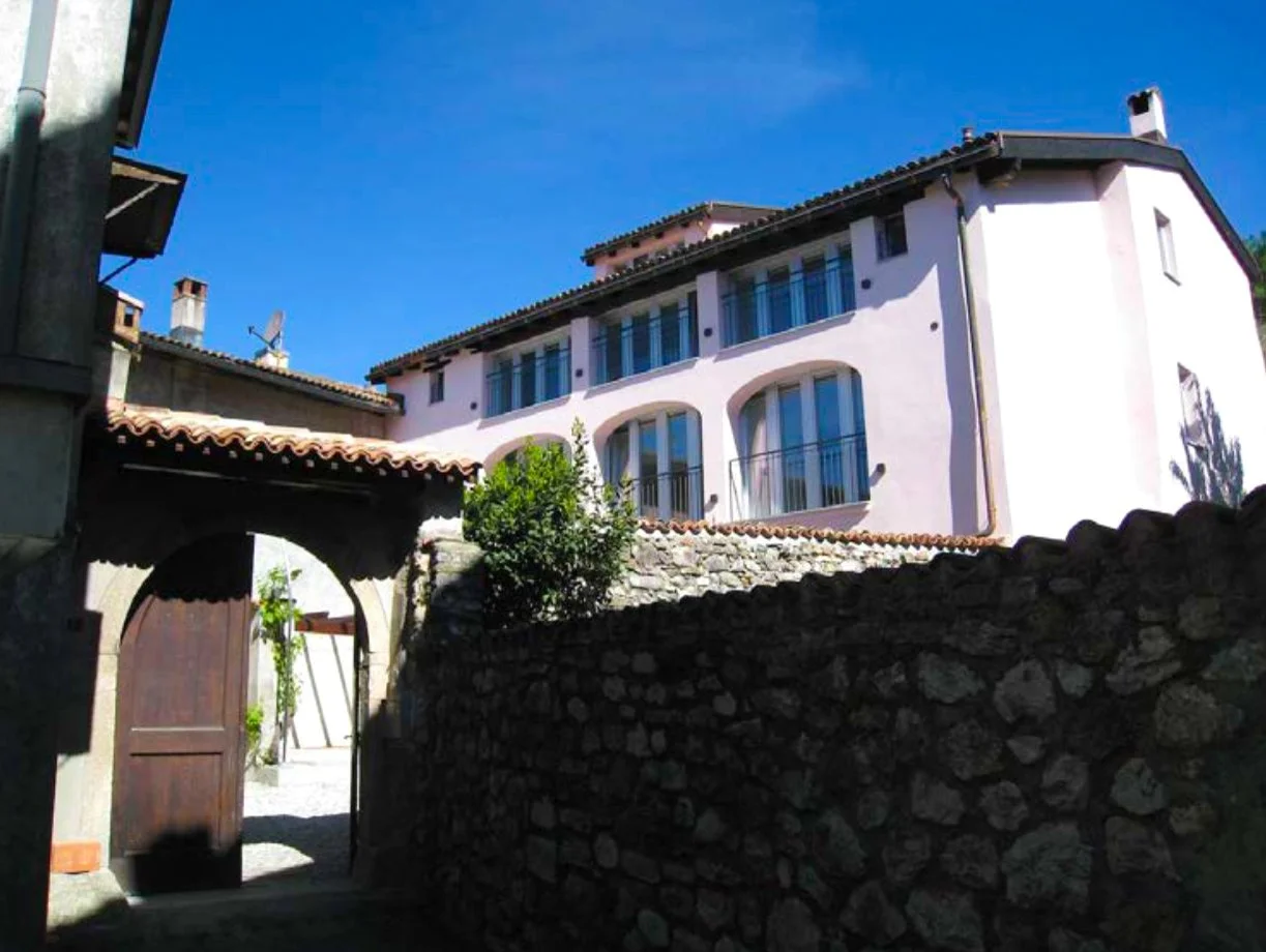 View of a multi-story house with white walls and large windows, surrounded by a stone wall and an archway entrance, under a clear blue sky.