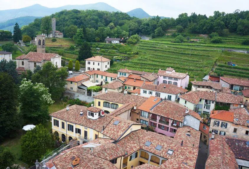 A scenic view of a village with colorful houses and terracotta roof tiles, surrounded by lush green trees and rolling hills in the background.