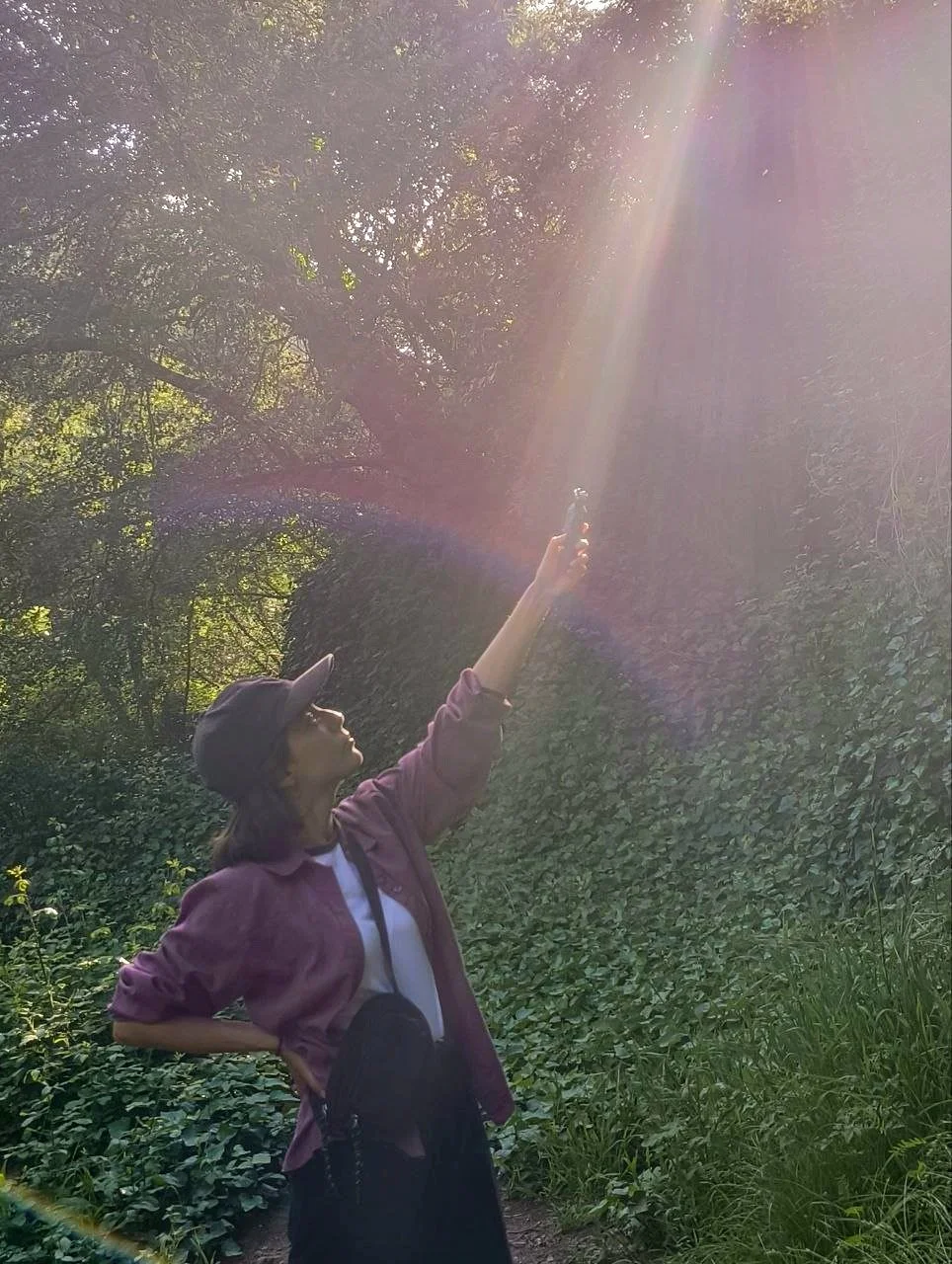 A person standing in a lush green forest, reaching up towards a beam of sunlight shining through the trees, wearing a sun hat, a jacket, and carrying a crossbody bag.