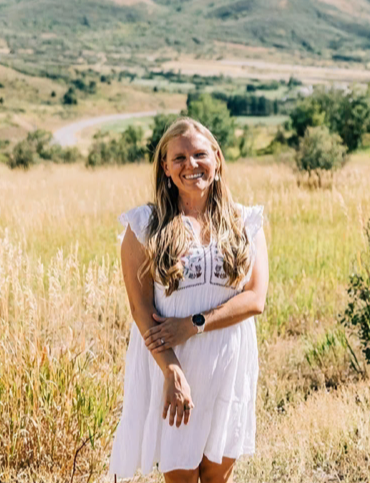 A woman in a white dress standing in a field with green hills and trees in the background, smiling at the camera.