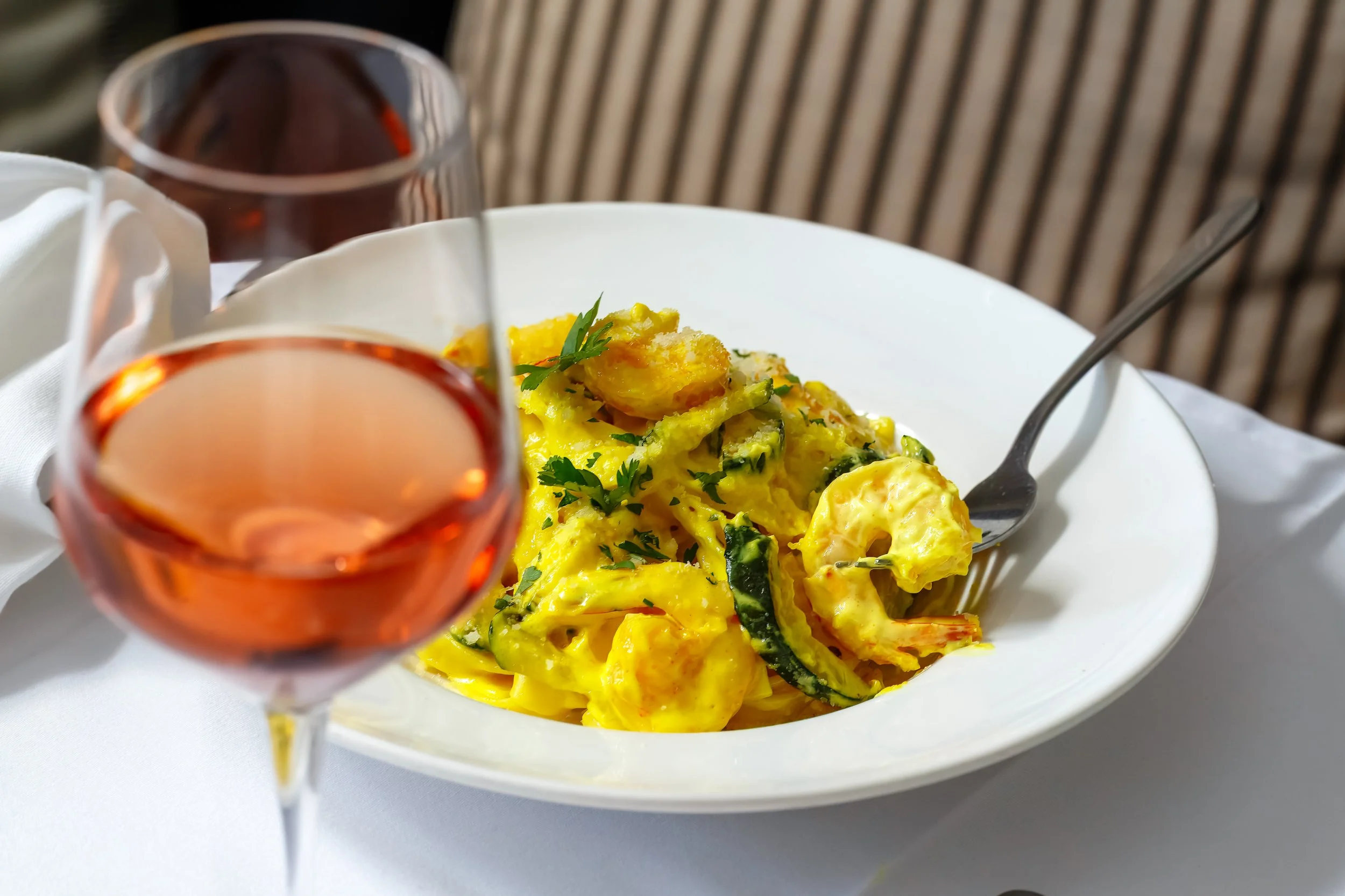A glass of rosé wine, a bowl of tagliatelle with courgette and prawns, and a fork on a white tablecloth.