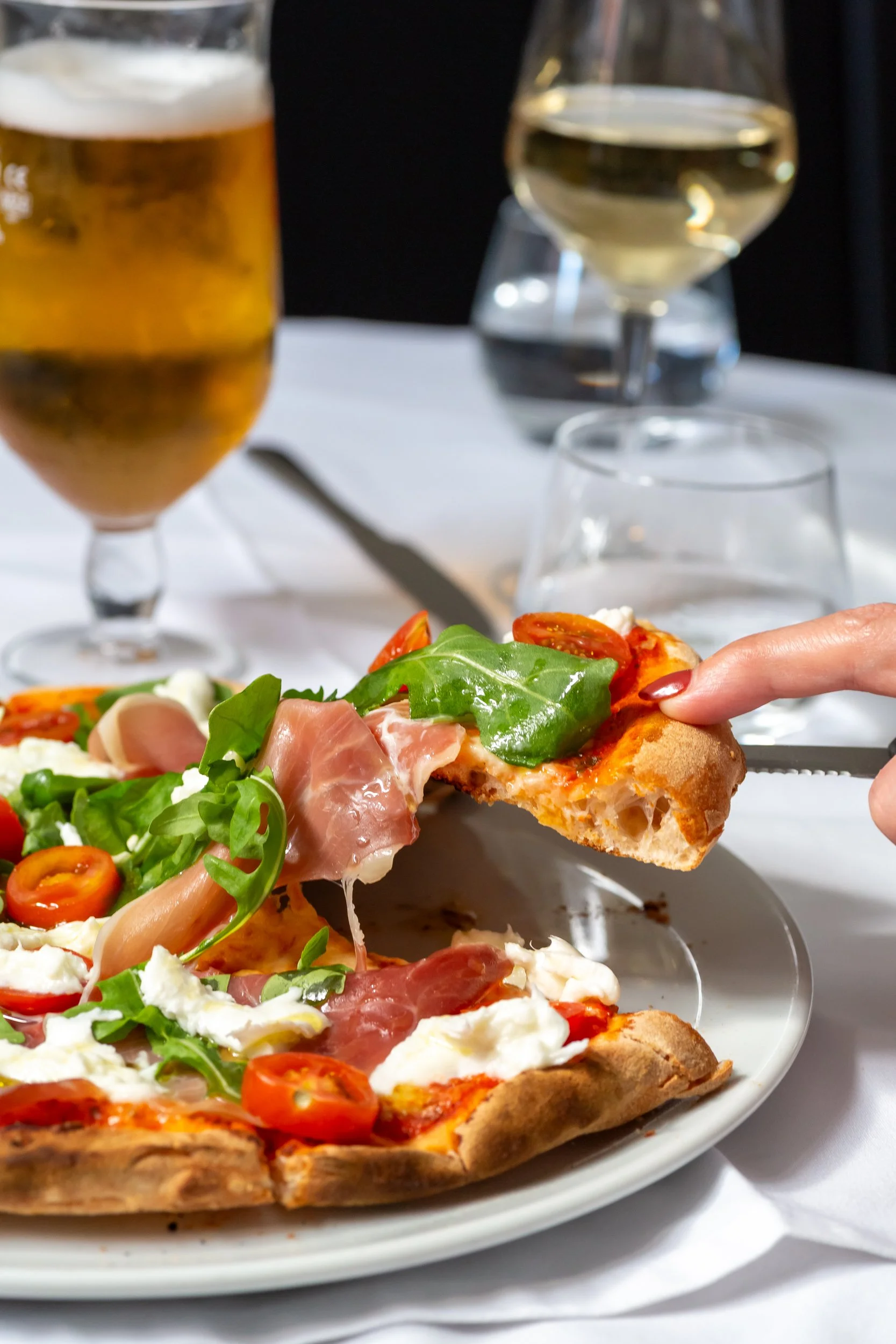 Close-up of a person holding a slice of Bufalina pizza  with toppings including cherry tomatoes, basil, and prosciutto, with a glass of white wine and a glass of beer in the background on a white table.
