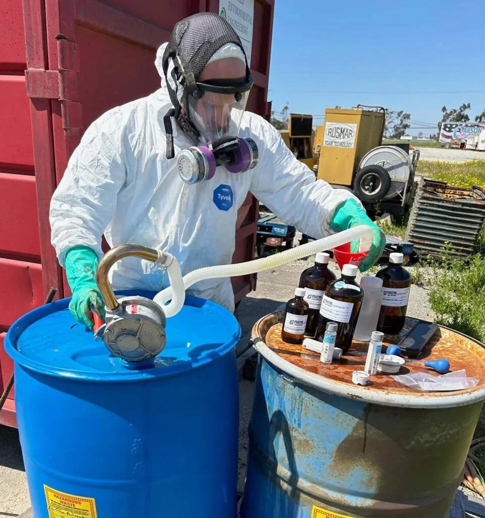 Person in a hazmat suit with drums and a funnel. There are chemical bottles near him.