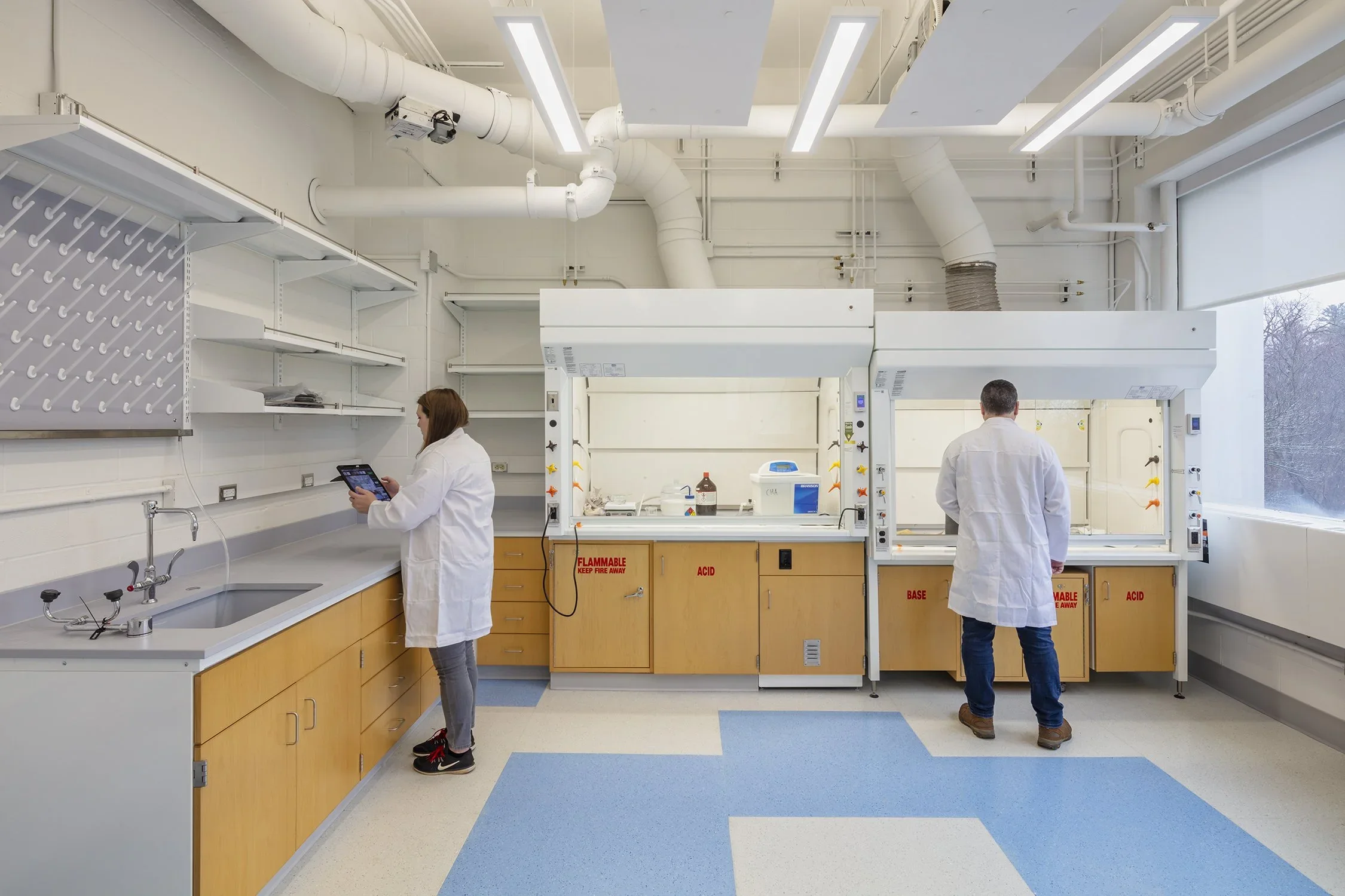 Two scientists in white lab coats working in a laboratory with fume hoods, shelves, and specialized equipment.