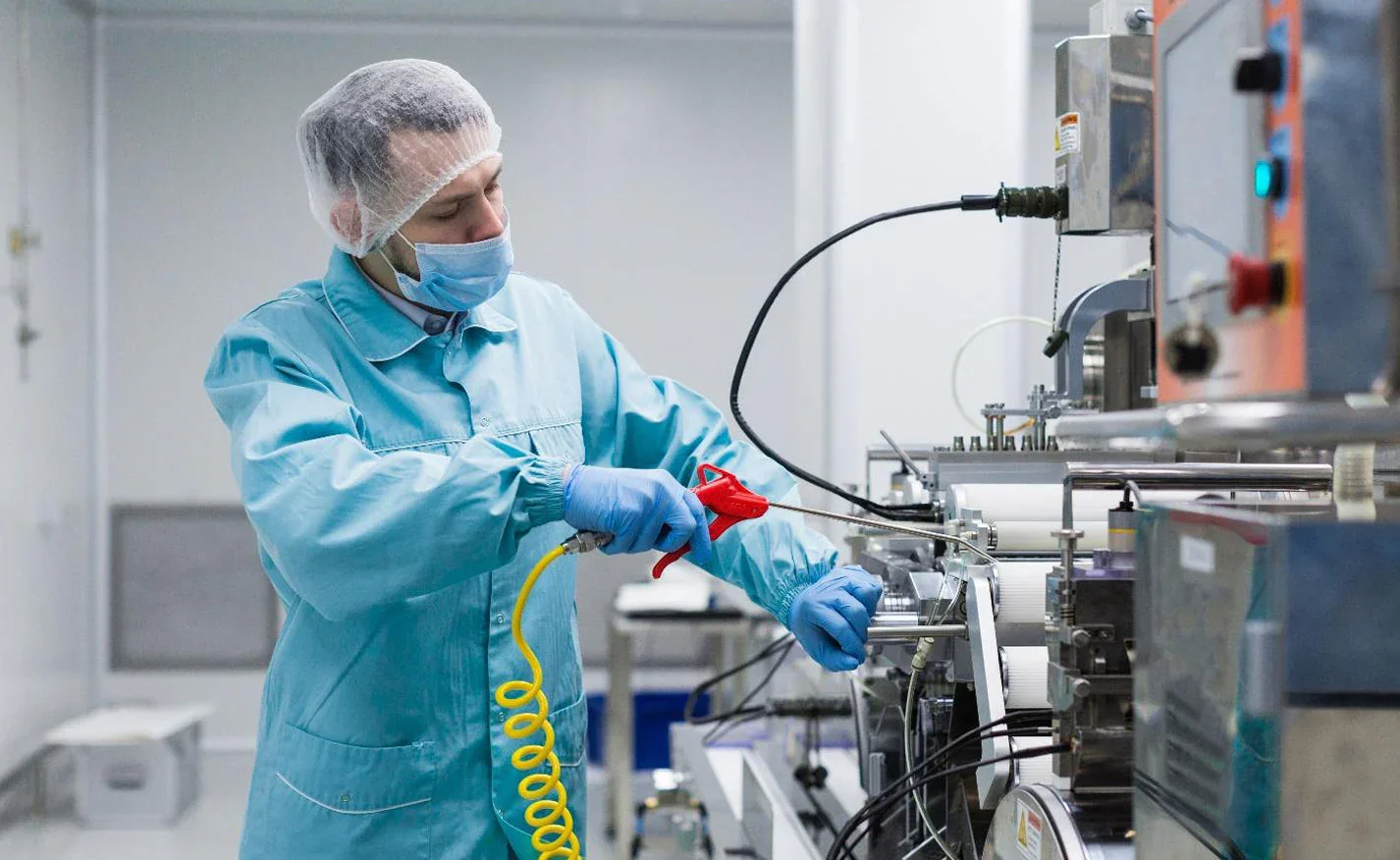 A scientist working in a laboratory, wearing a hairnet, face mask, and gloves, using a tool on industrial equipment.