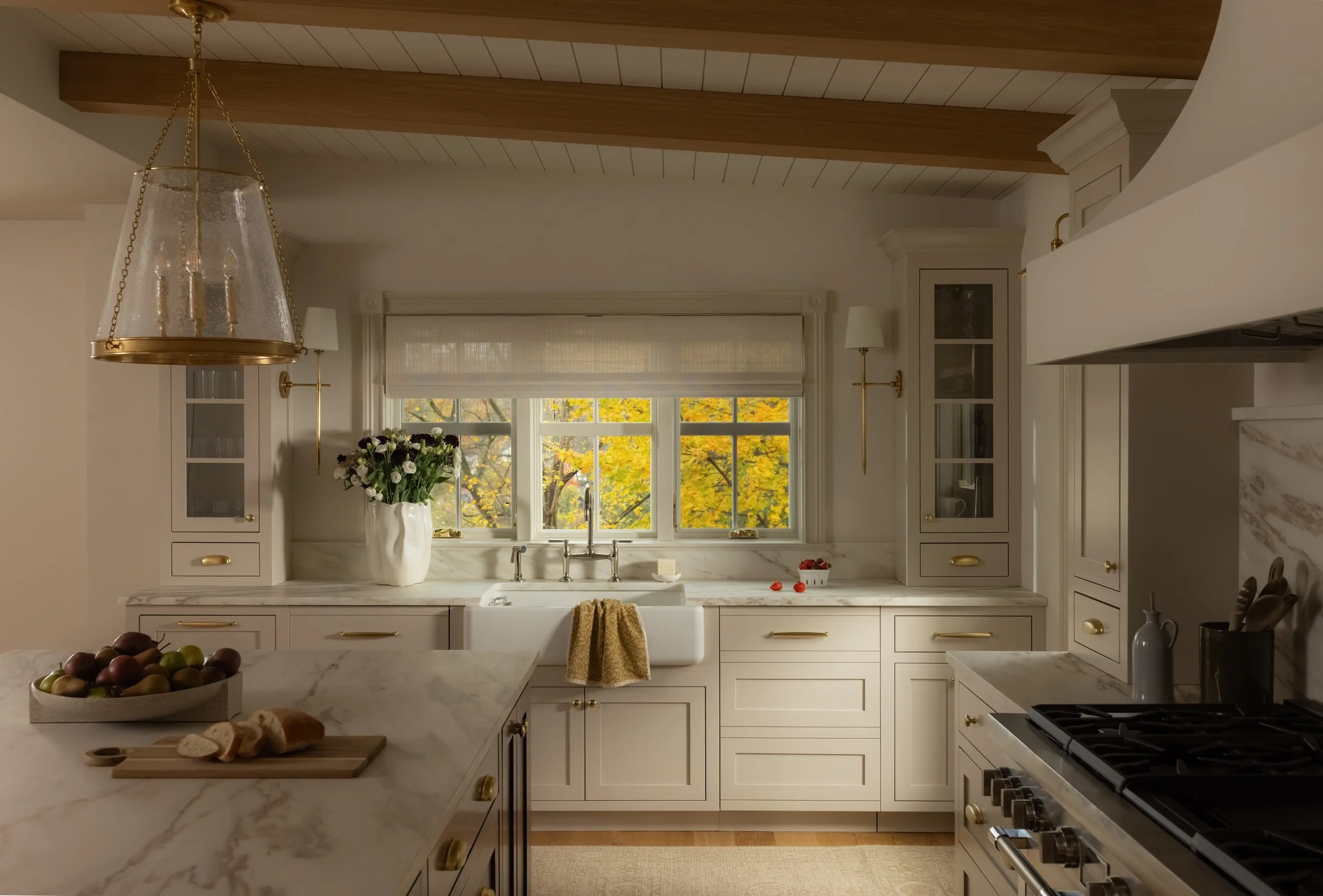 Warm, neutral kitchen featuring architectural wood beams and natural light streaming from a window, with a view of autumn trees in Roslyn, NY. Photographed by Raquel Langworthy