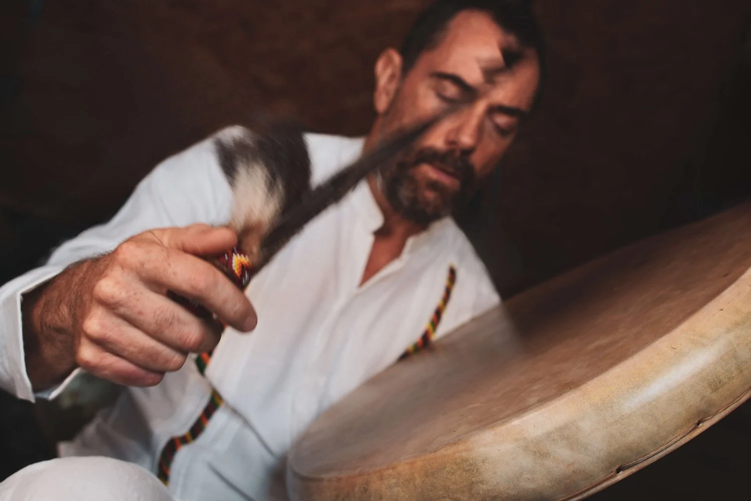 A man with dark hair and a beard, dressed in white shirt and trousers, playing a large shamanic drum with his eyes closed. He has a  beater and feathers in his hand which he's playing the drum with..