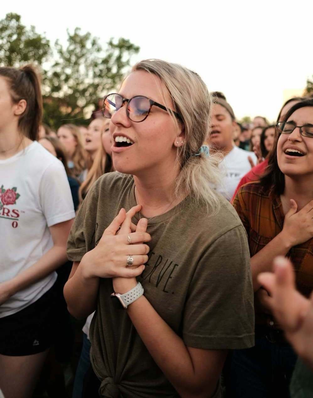 A young woman singing soulfully with her hands joined together resting upon her heart. Surrounding this woman is other women also singing with their hands resting upon their hearts too.