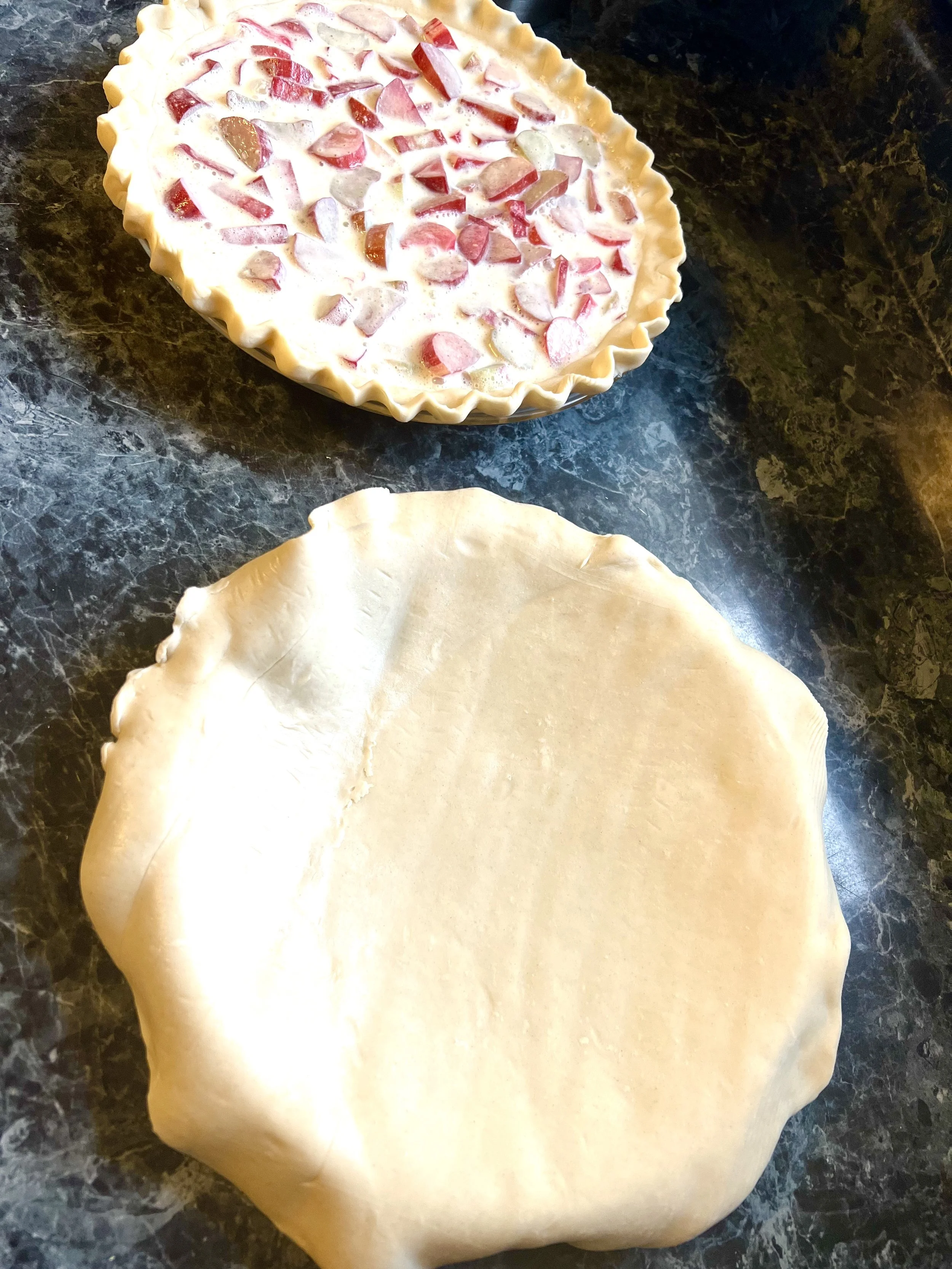 A partially assembled pie on a dark, green marble surface. The pie crust is rolled out and shaped in a circular form. The pie top with a fluted edge is filled with a fruit filling, likely with chunks of apples or similar fruit, and appears ready to be covered with the crust for baking.