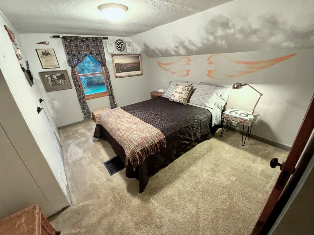 A bedroom with a bed covered in a black blanket, decorated with pillows, and a throw blanket at the foot. A small side table with a lamp and a sign reading '66' is next to the bed. The wall has a painted design of two stylized, orange and white flames. A window with patterned curtains is on the back wall, and framed pictures and wall decorations are also visible.
