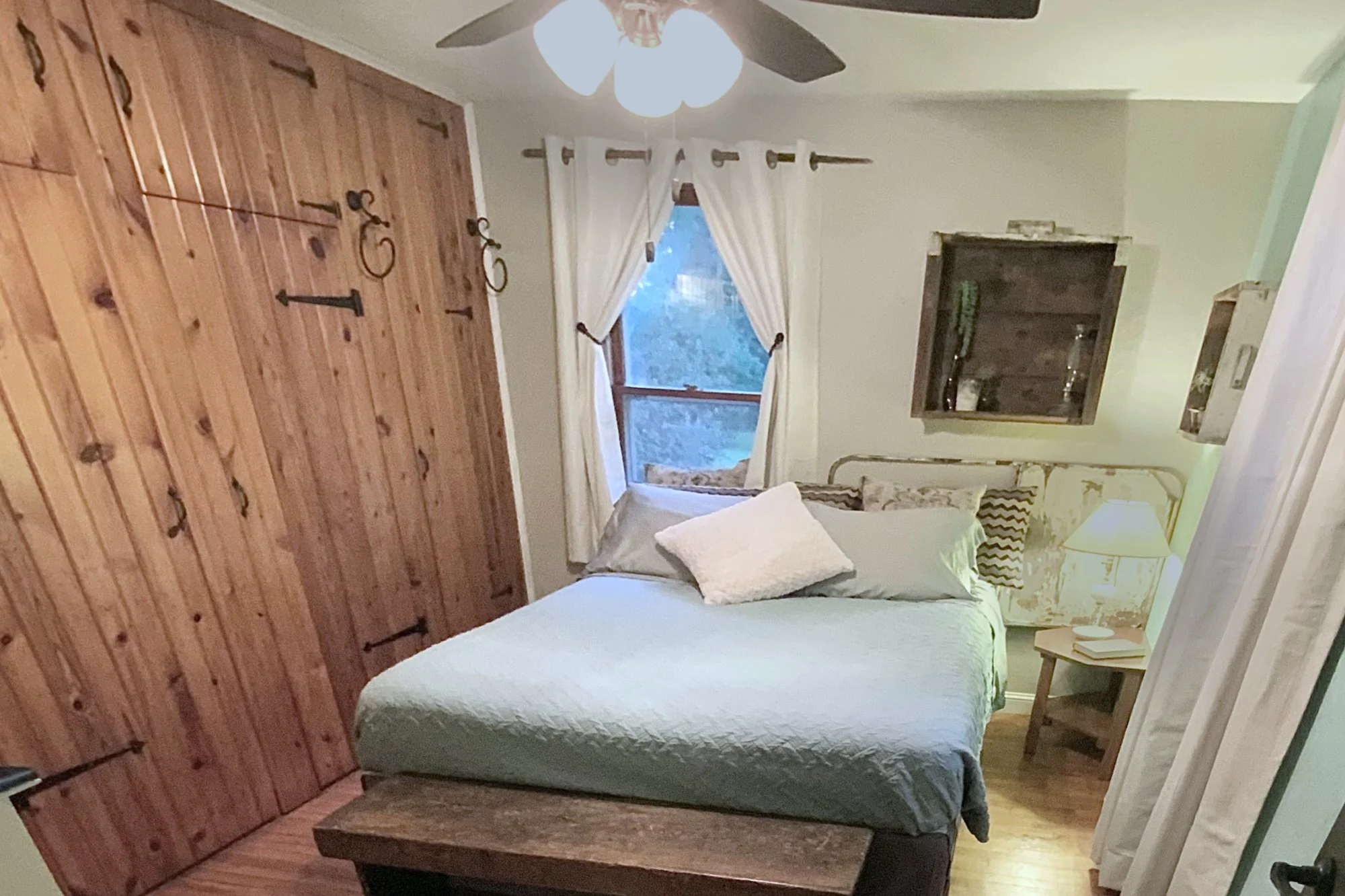 A cozy bedroom with a wooden wall on the left, white curtains over a window, a bed with a light-colored quilt and pillows, a nightstand with a lamp, and a mounted wooden display shelf.