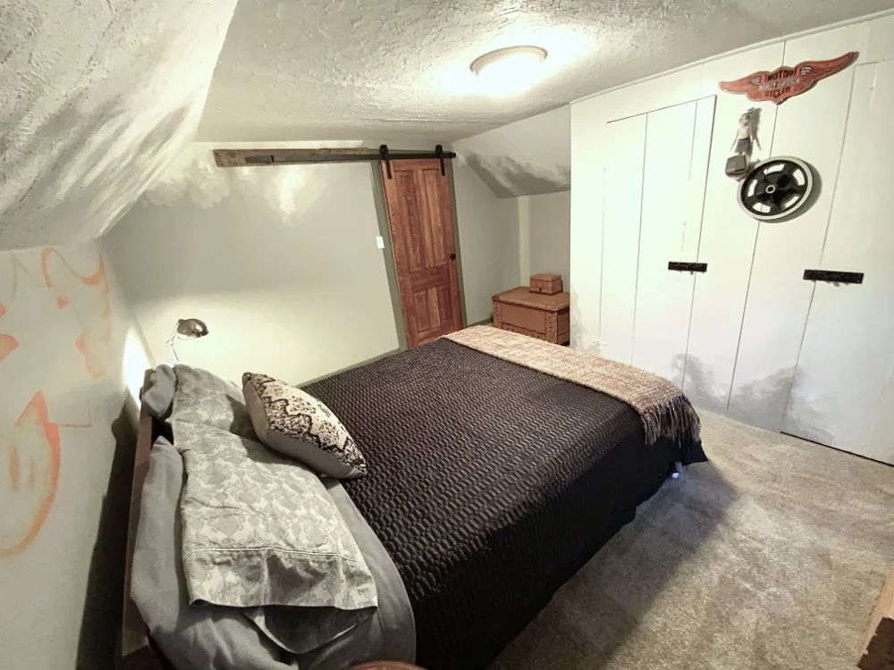 A small bedroom with a bed covered in a black textured blanket, grey pillows, and a decorative pillow. There is a wooden dresser and a sliding barn door on a metal track. The wall on the right has white informal closet doors, with a Harley-Davidson sign and a steering wheel decoration hanging above.