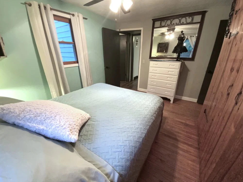 A cozy bedroom with a bed covered in light green bedding, a plush pillow, a wooden wardrobe, a white dresser with a mirror, a window with white curtains, and hardwood floors.