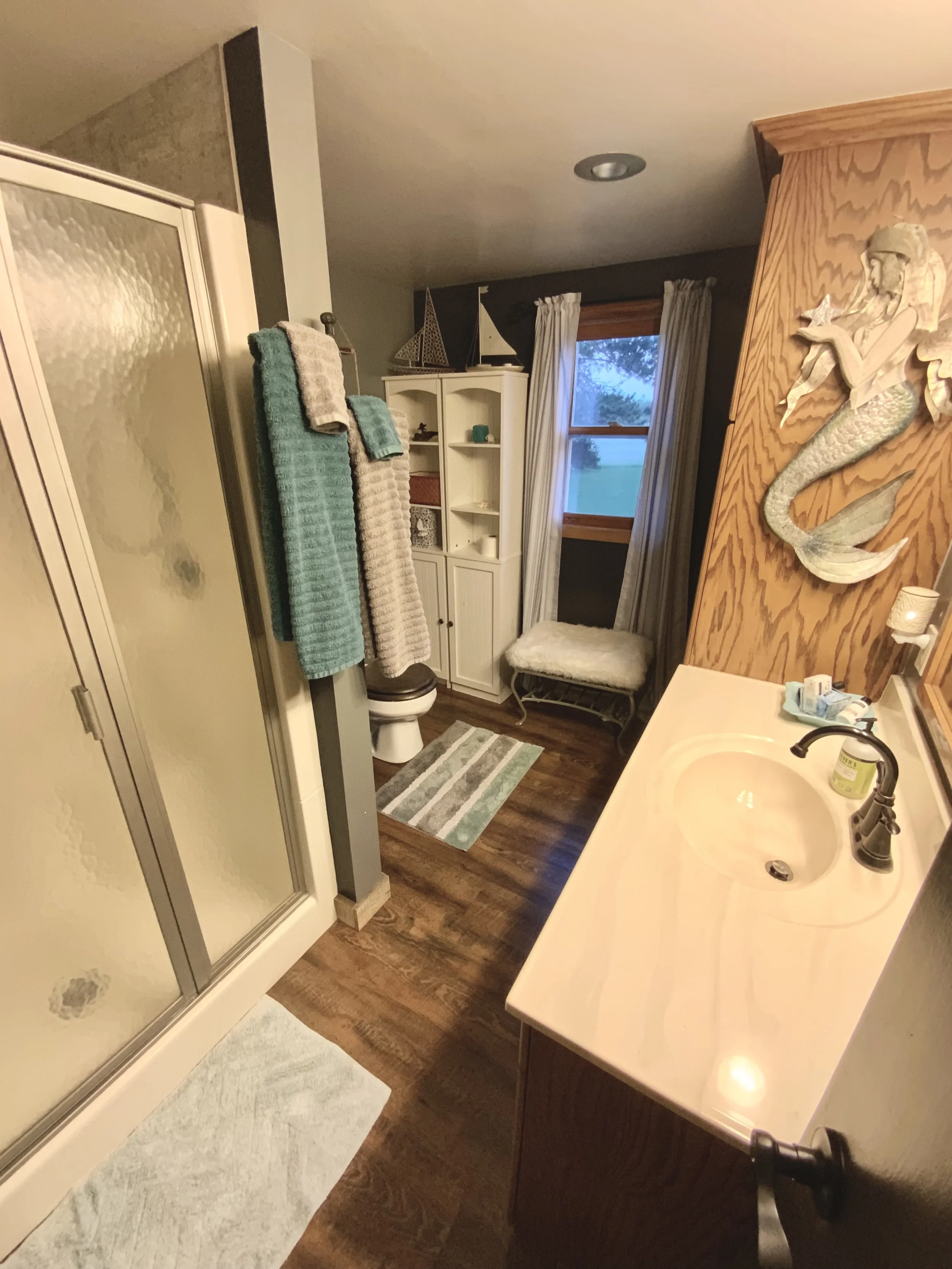A bathroom with a shower stall on the left, towels hanging on a rack, a white cabinet with shelves, a window with curtains, a cushioned bench, a sink with a countertop, a decorative mermaid wall sculpture, and wooden flooring.