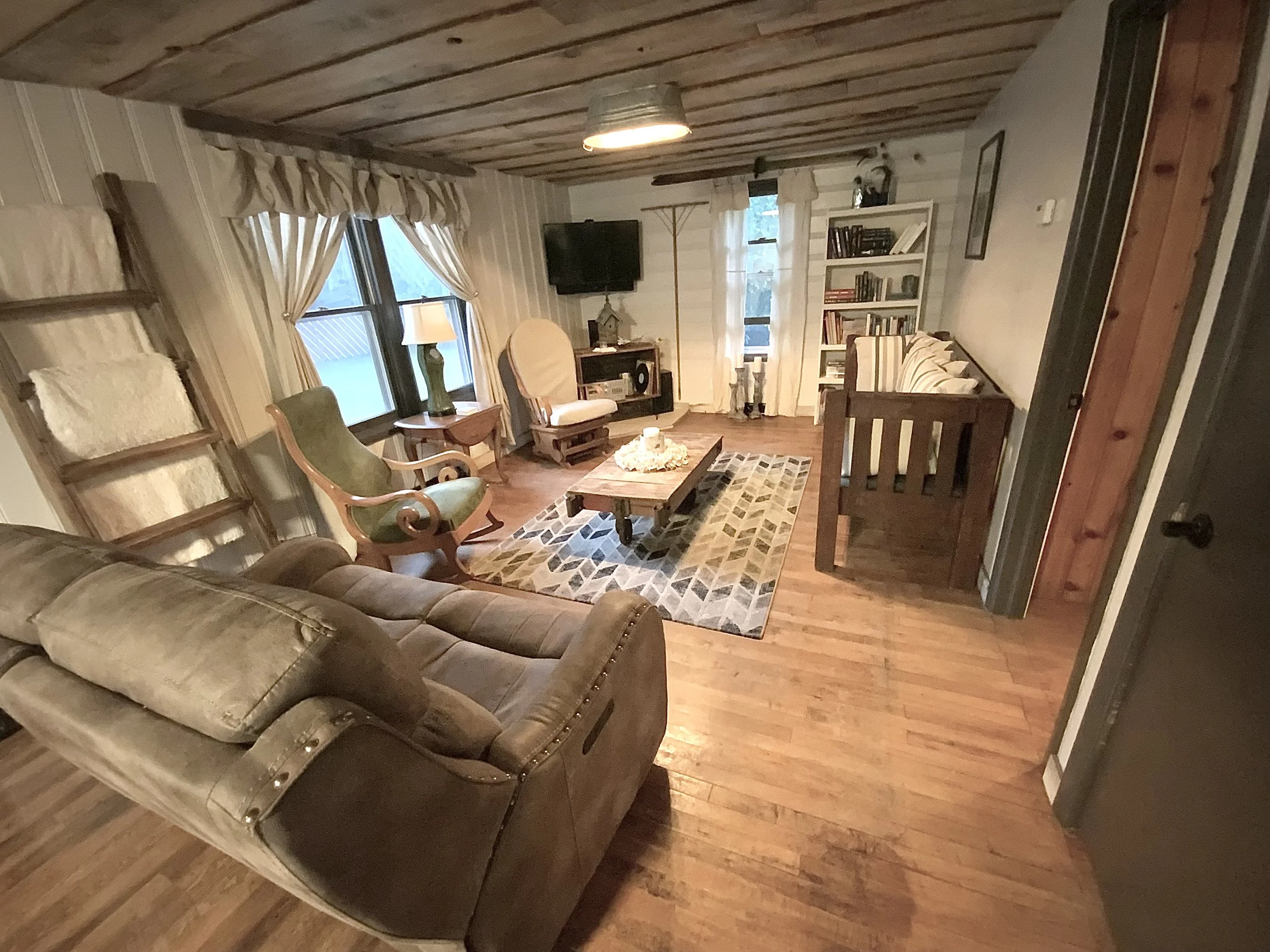 A cozy living room with a wooden ceiling, hardwood floors, a brown leather sofa, a green armchair, a white armchair, a coffee table on a geometric patterned rug, a bookshelf, a TV, and windows with curtains.