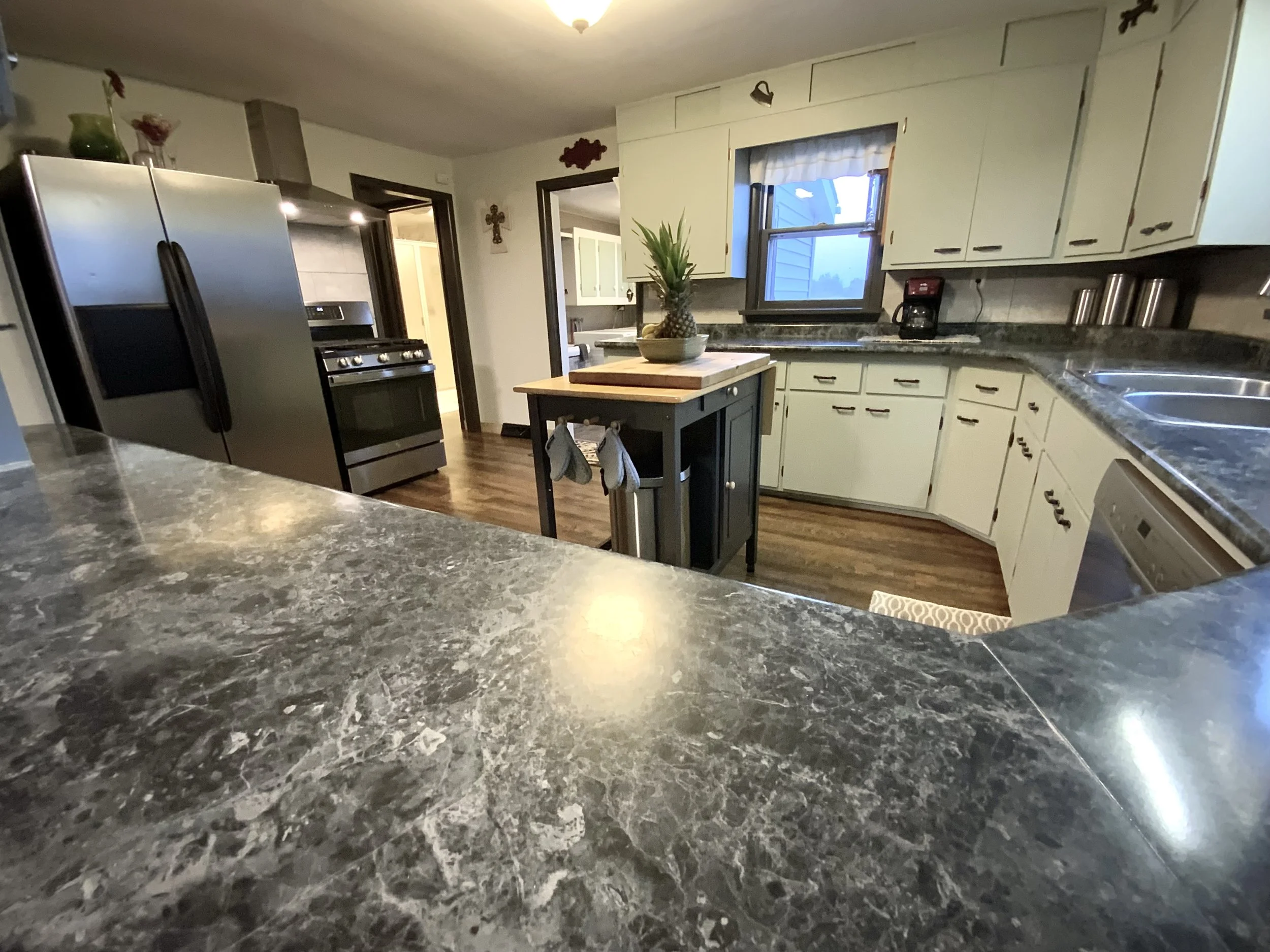 A kitchen with silvermist cabinets, a stainless steel refrigerator, a dark-colored kitchen island with a pineapple on top, a stove, a window above the sink, a coffee maker, and wooden flooring. There is a doorway leading to another room.