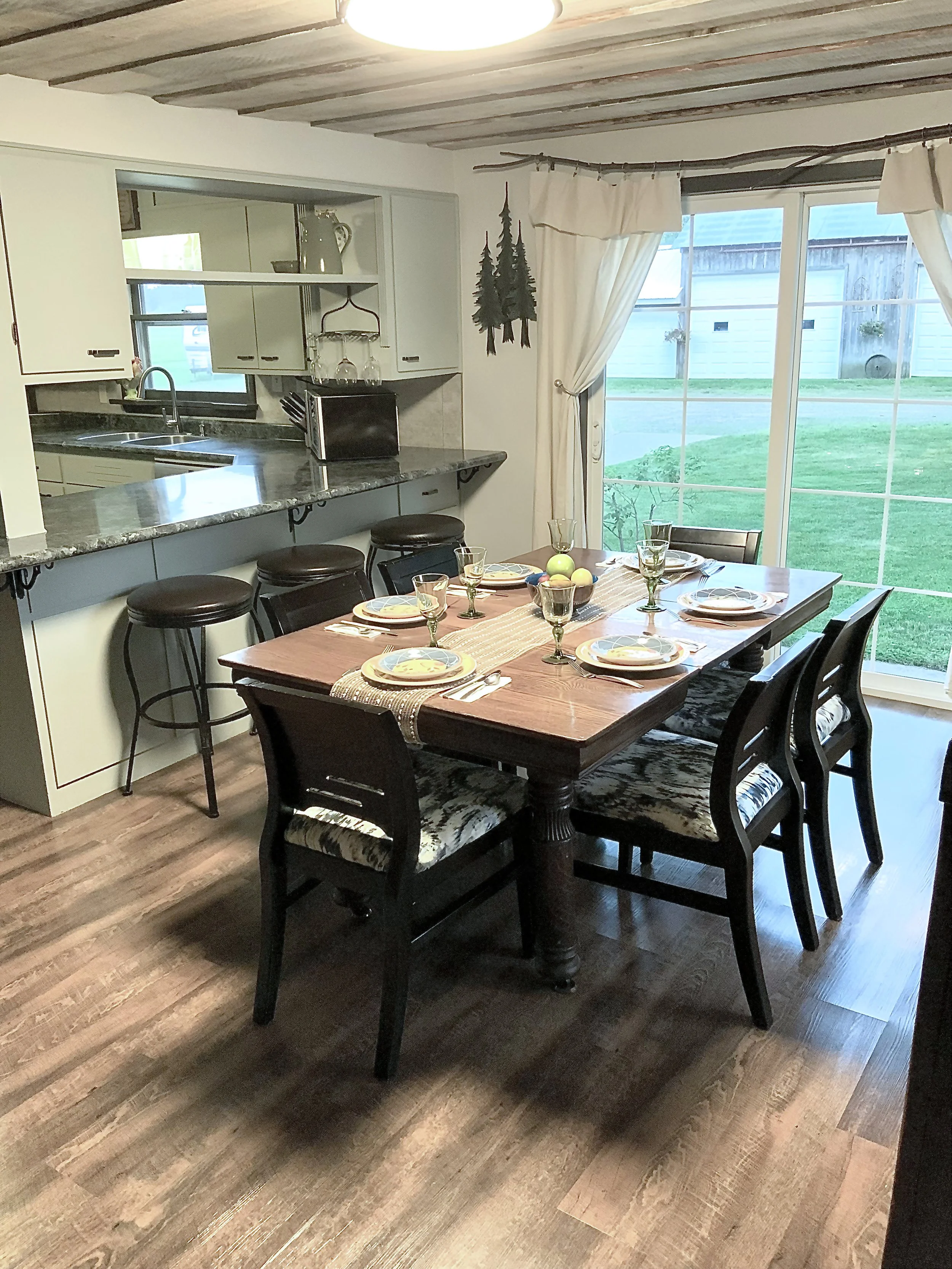 A dining room with a wooden table set for six, with plates, glasses, and a centerpiece of apples. There is a large window showing a green yard outside. A kitchen counter with bar stools is behind the table, and the room has a rustic ceiling with wooden beams.