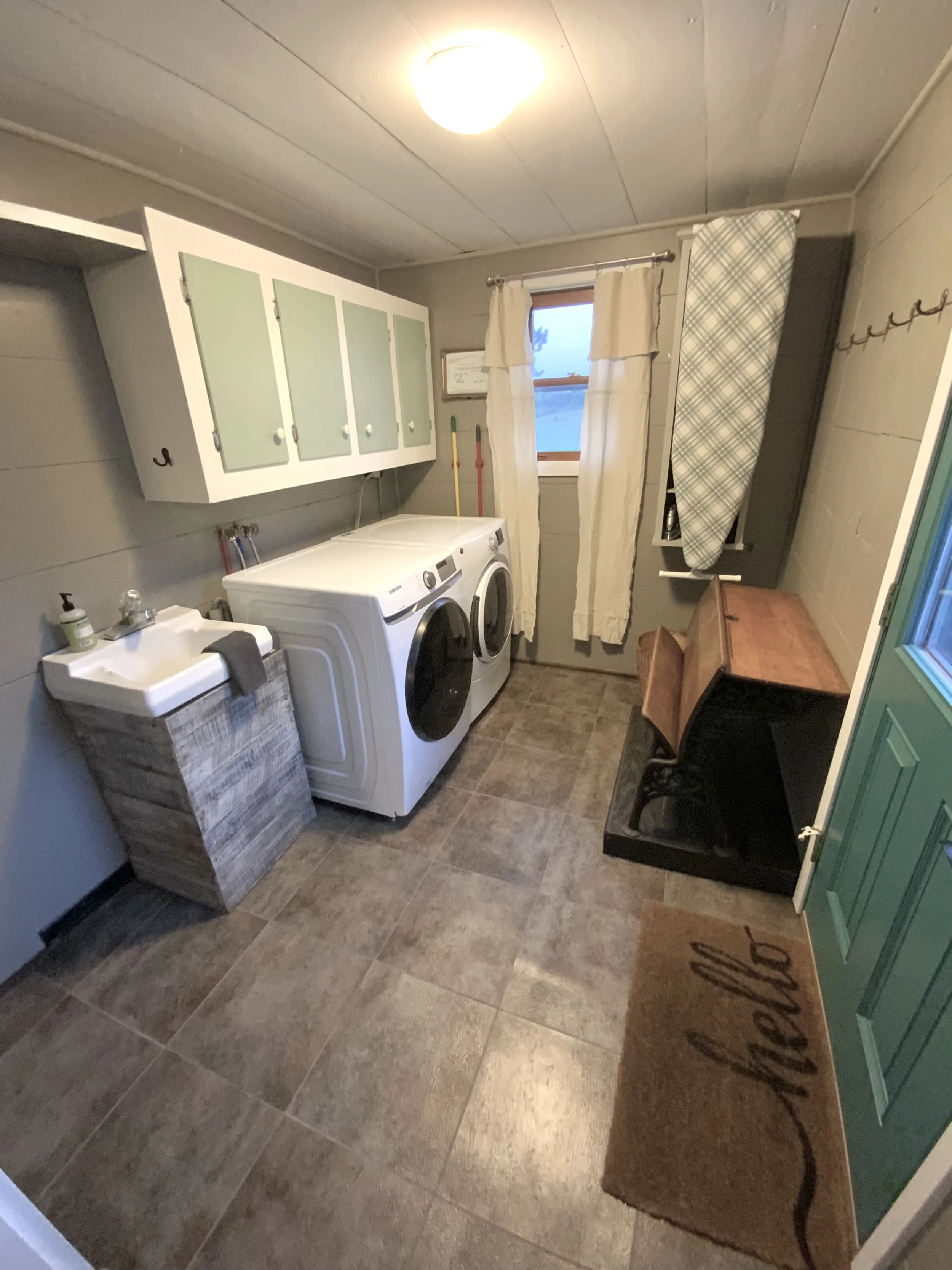 A laundry room with a washing machine and dryer, window with curtains, green cabinet, small sink, ironing board, wooden desk, and a welcome mat.