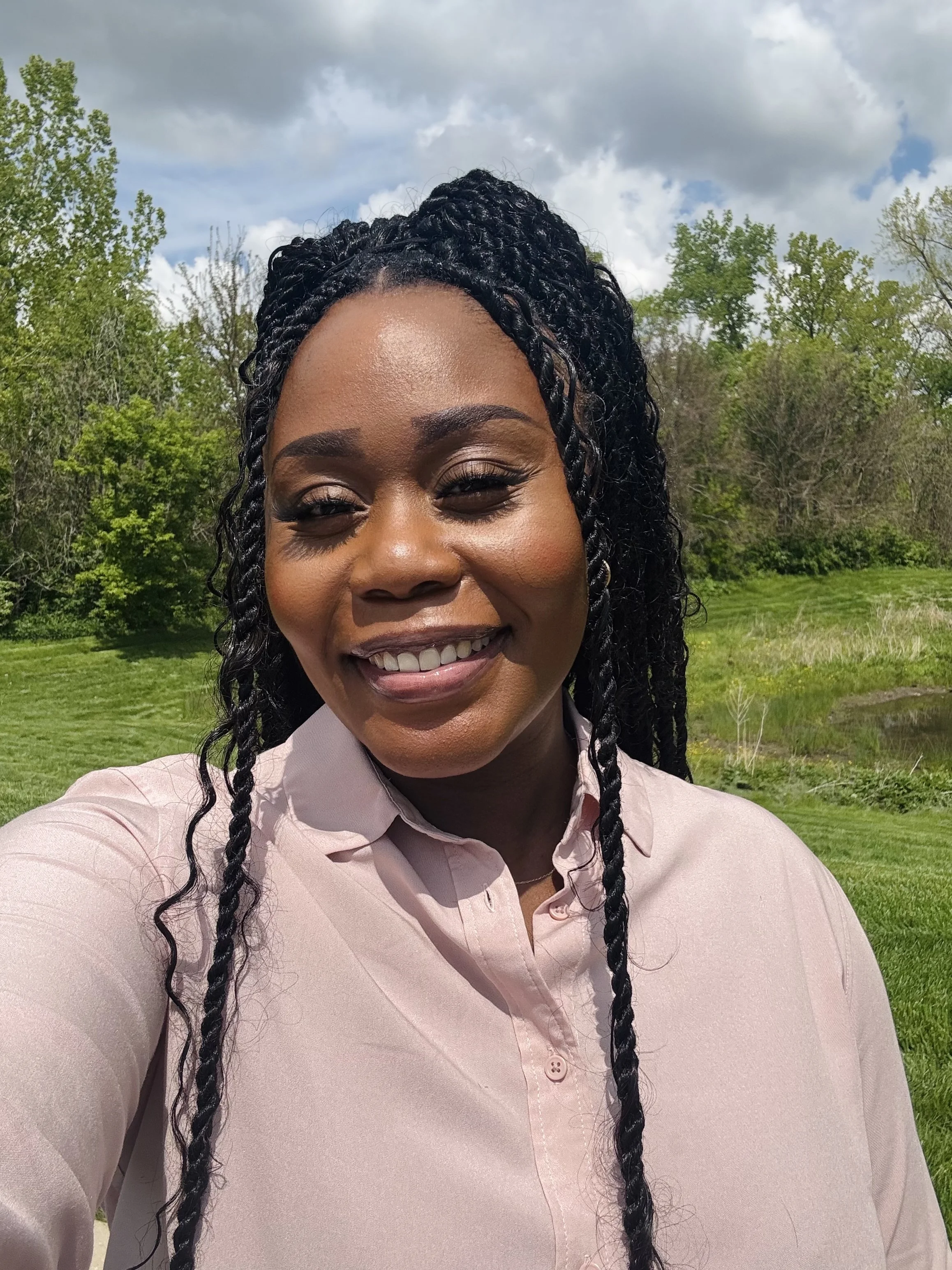 Close-up of a smiling African American woman with braided hair, wearing a light pink blouse, outdoors with green trees and cloudy sky in the background.