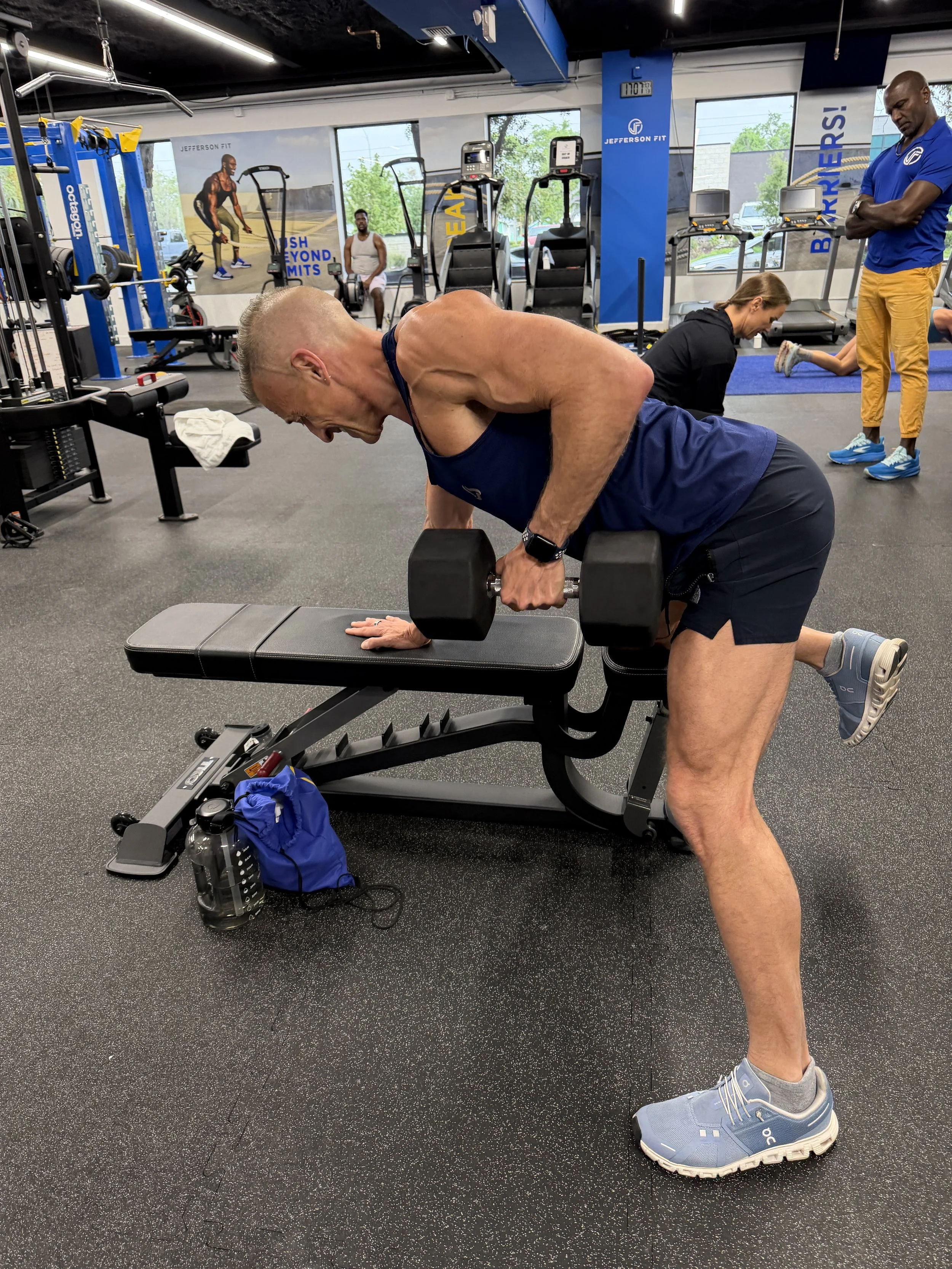 Member performing a dumbbell row at Jefferson Fit in downtown Sarasota, with a trainer assisting another member in the background.