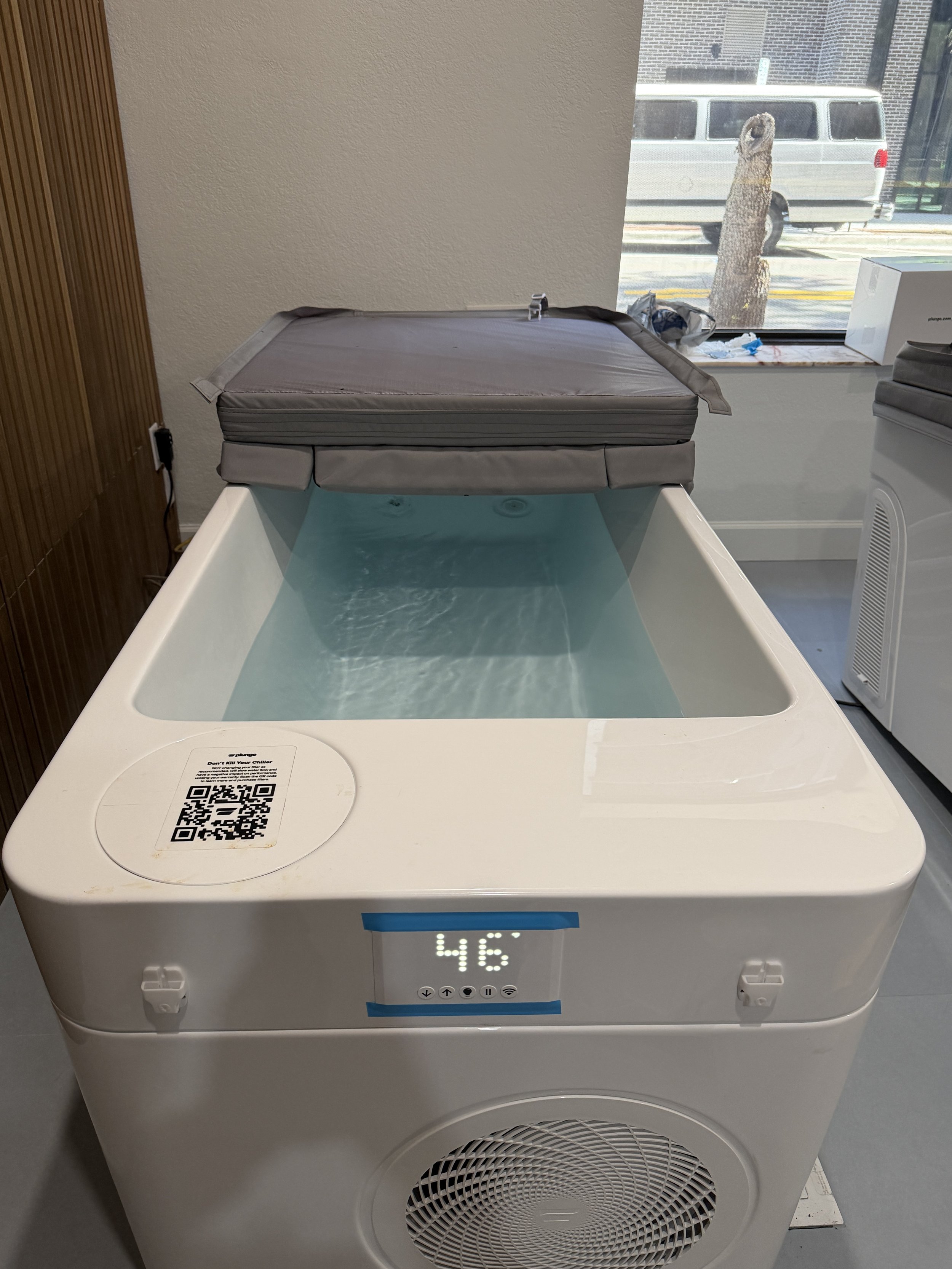 A laundry or drying machine with a built-in water bath, with a digital display showing 46 degrees and a gray cover on top, located inside a room near a window.