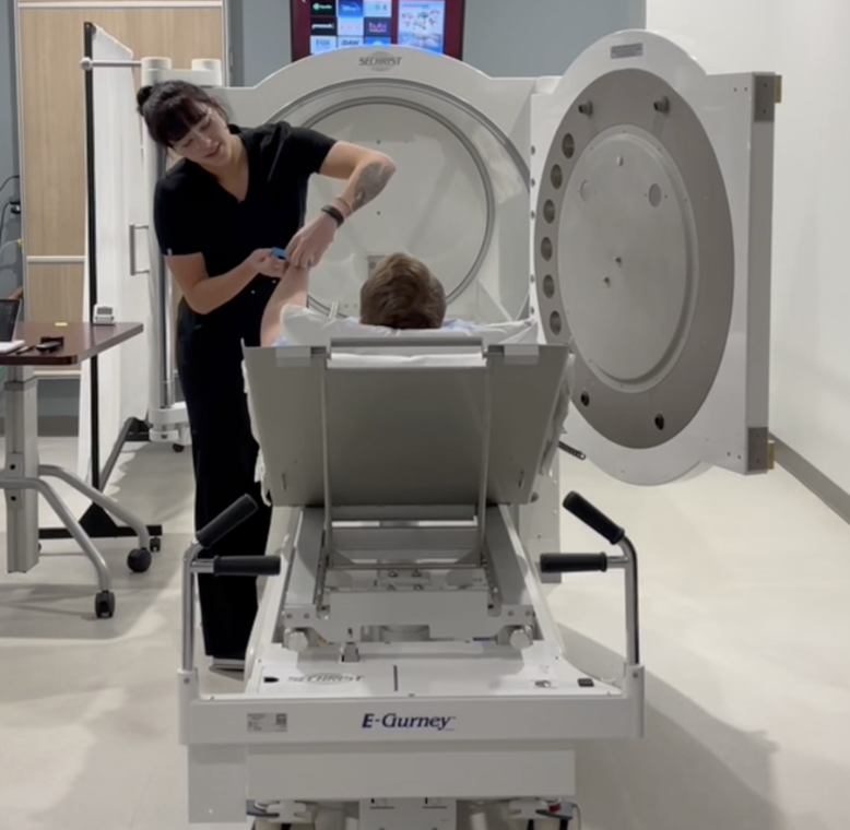 A person lying in front of a hyperbaric chamber, while a technician prepares them by adjusting their grounding strap, in a medical facility.