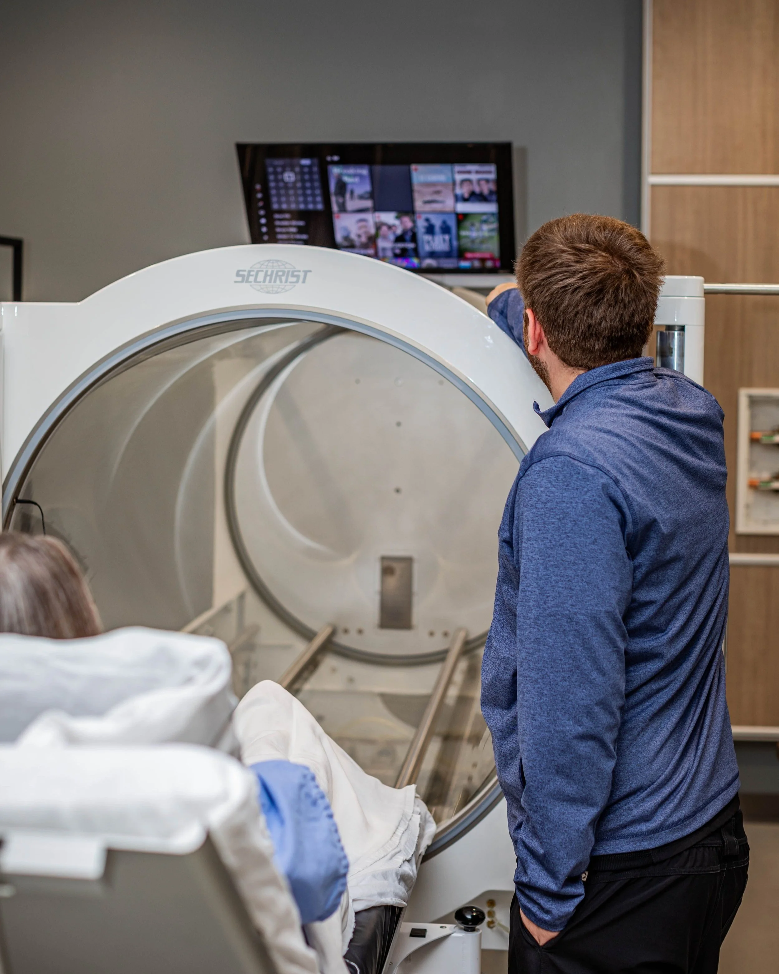 A person with a blue shirt and short hair standing next to a patient lying on a medical bed, preparing for hyperbaric treatment, with a tv above the chamber for the patient to watch during treatment.