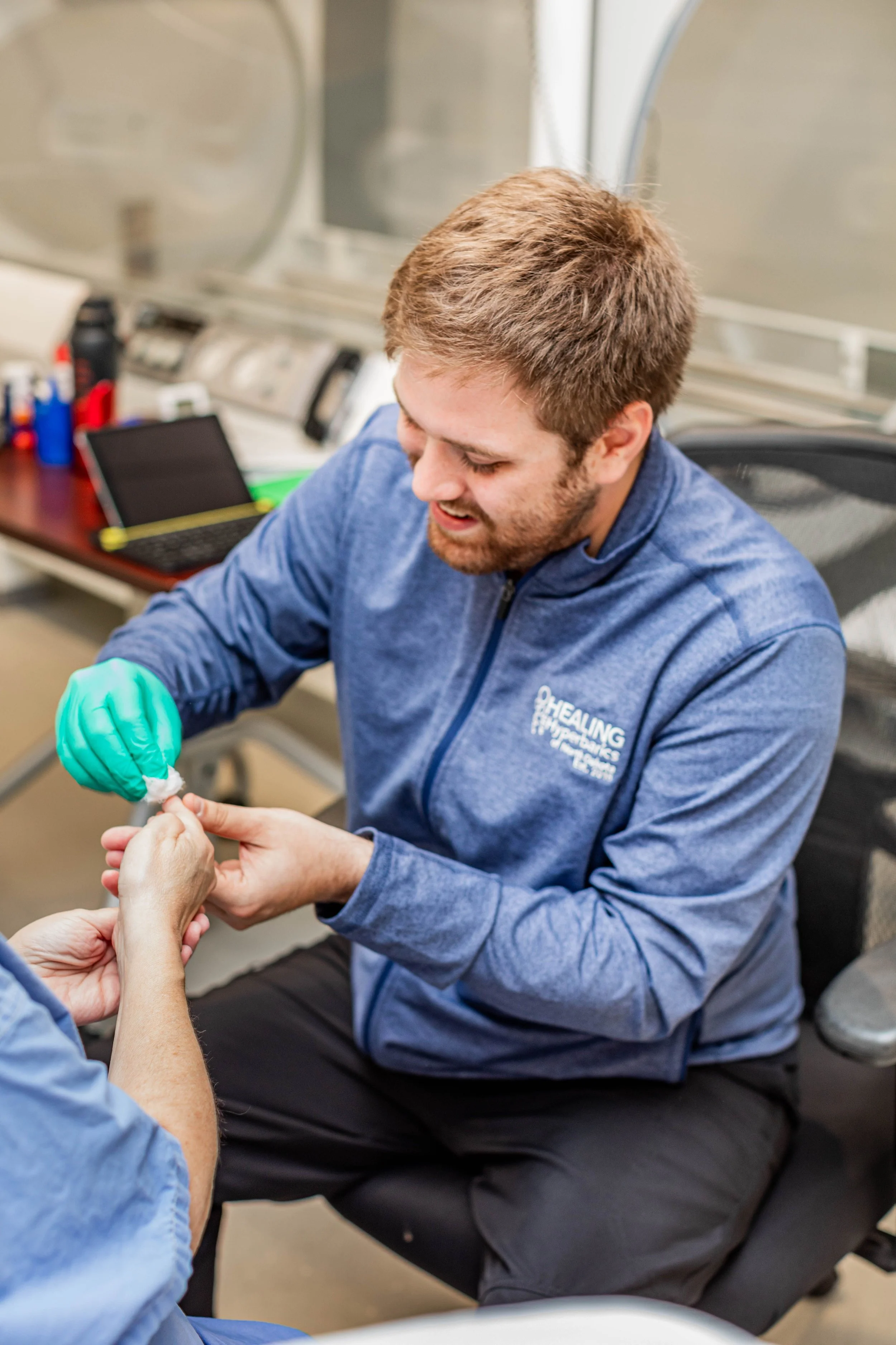 A man dressed in a blue jacket with a logo on it is performing a blood glucose test for a patient before a treatment.