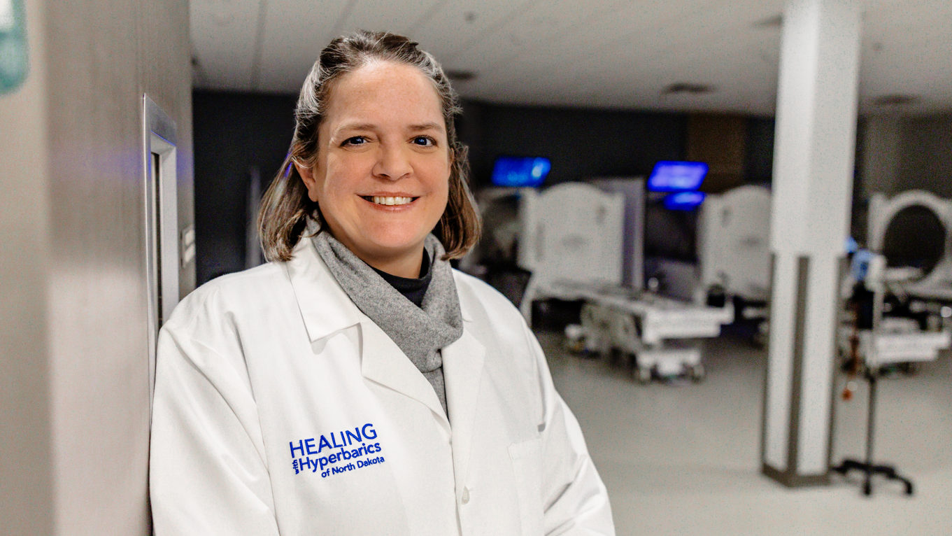 Dr. Daphne Denham, MD, FACS, FACCWS, standing in the Healing with Hyperbarics of North Dakota clinic in front of hyperbaric oxygen therapy chambers.