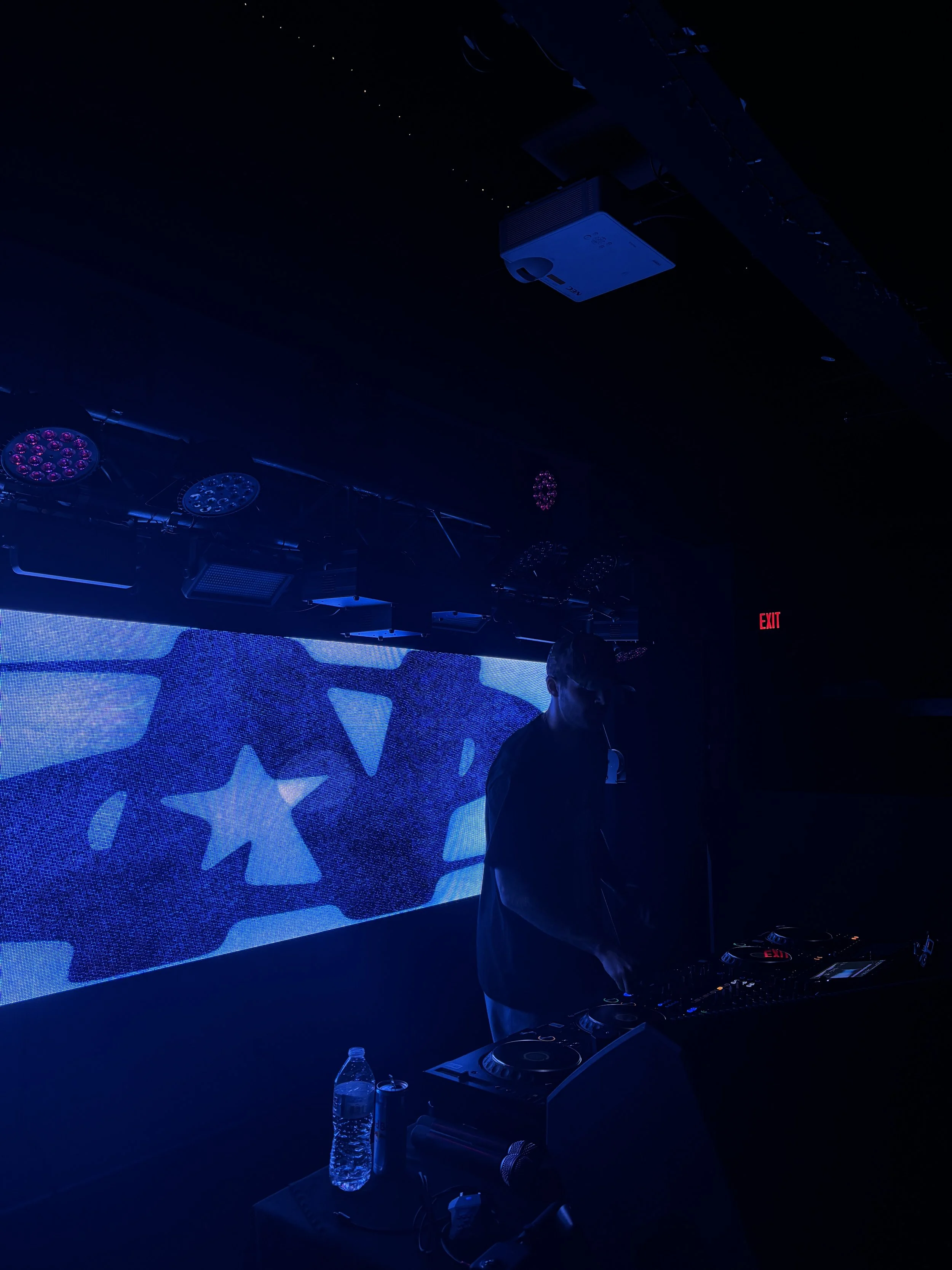 A DJ performing in a dark nightclub with blue lighting and a large LED screen displaying stars and a celestial design in the background. There are DJ equipment and a water bottle on a nearby table.