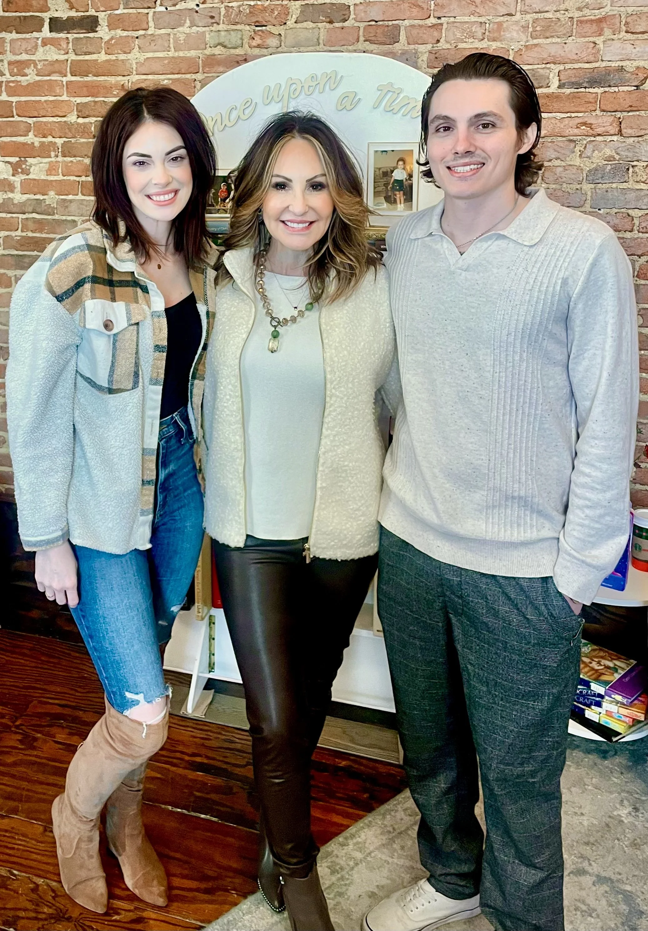 Three people standing close together, smiling, in front of a brick wall and a white sign with gold writing that says 'Once upon a time.'