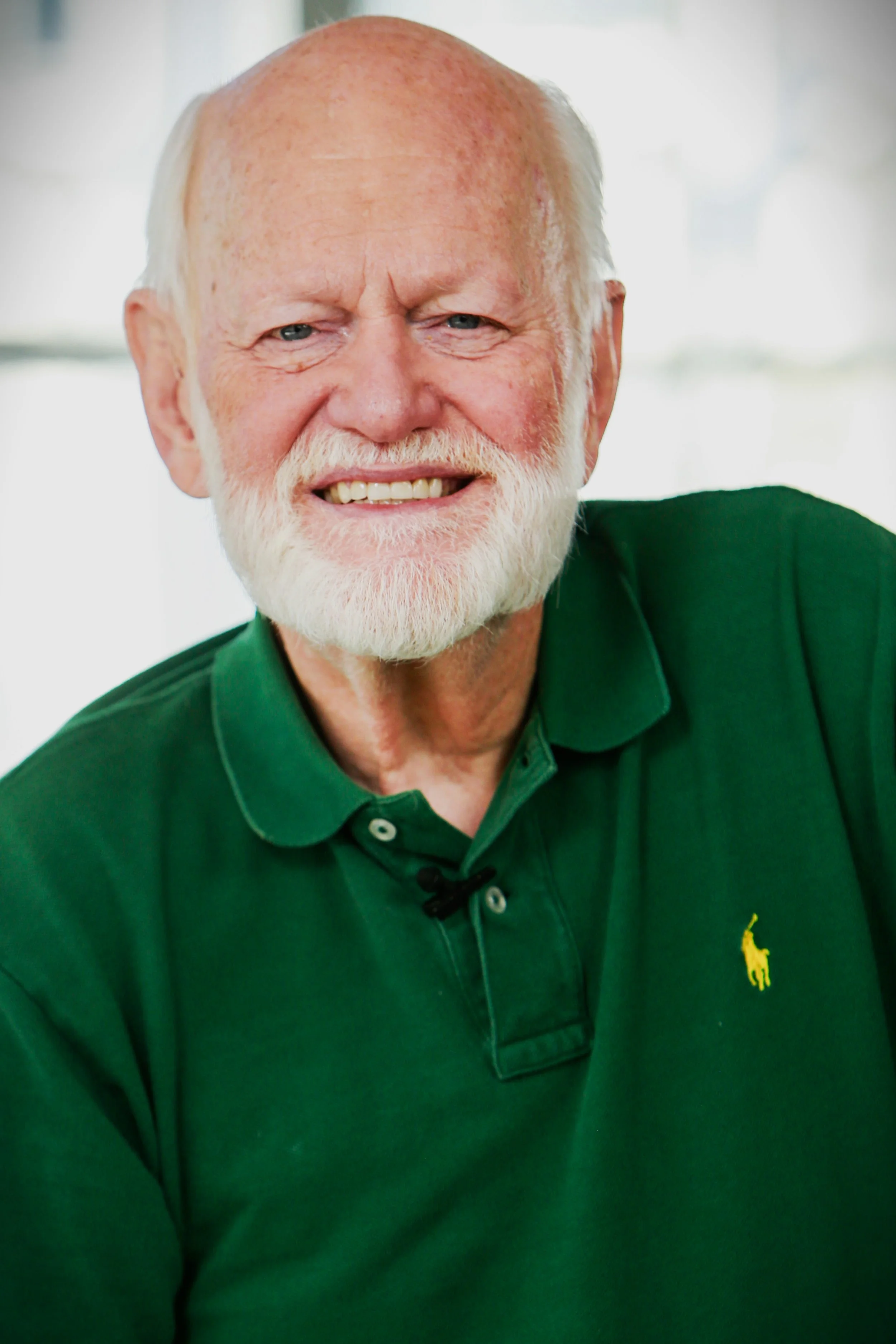 Close-up of an elderly man with a white beard and mustache, smiling, wearing a green polo shirt with a small yellow polo logo on the chest.