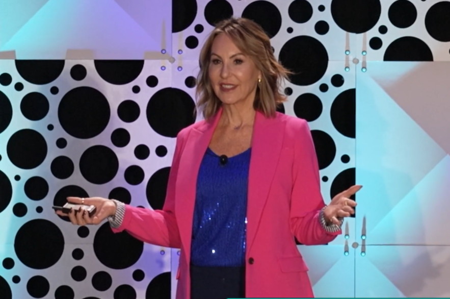 A woman with shoulder-length blonde hair wearing a bright pink blazer over a blue sequined top, standing against a black and white polka dot patterned background, gesturing with her hands while holding a remote control.