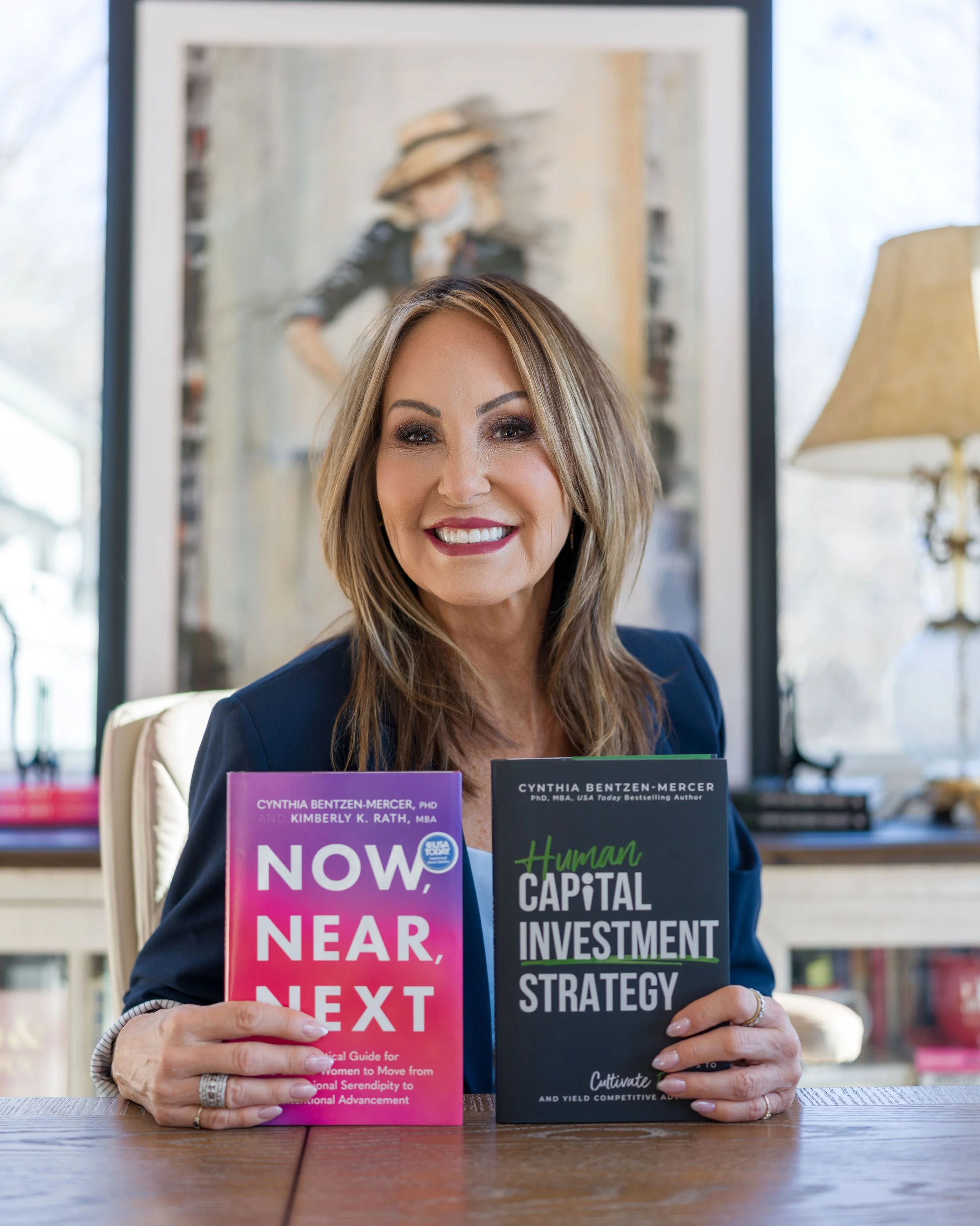 A woman with shoulder-length brown hair smiling at the camera, holding two books titled 'Now, Near, Next' and 'Human Capital Investment Strategy'. She is seated at a wooden table in a well-lit room with bookshelves and a lamp in the background.