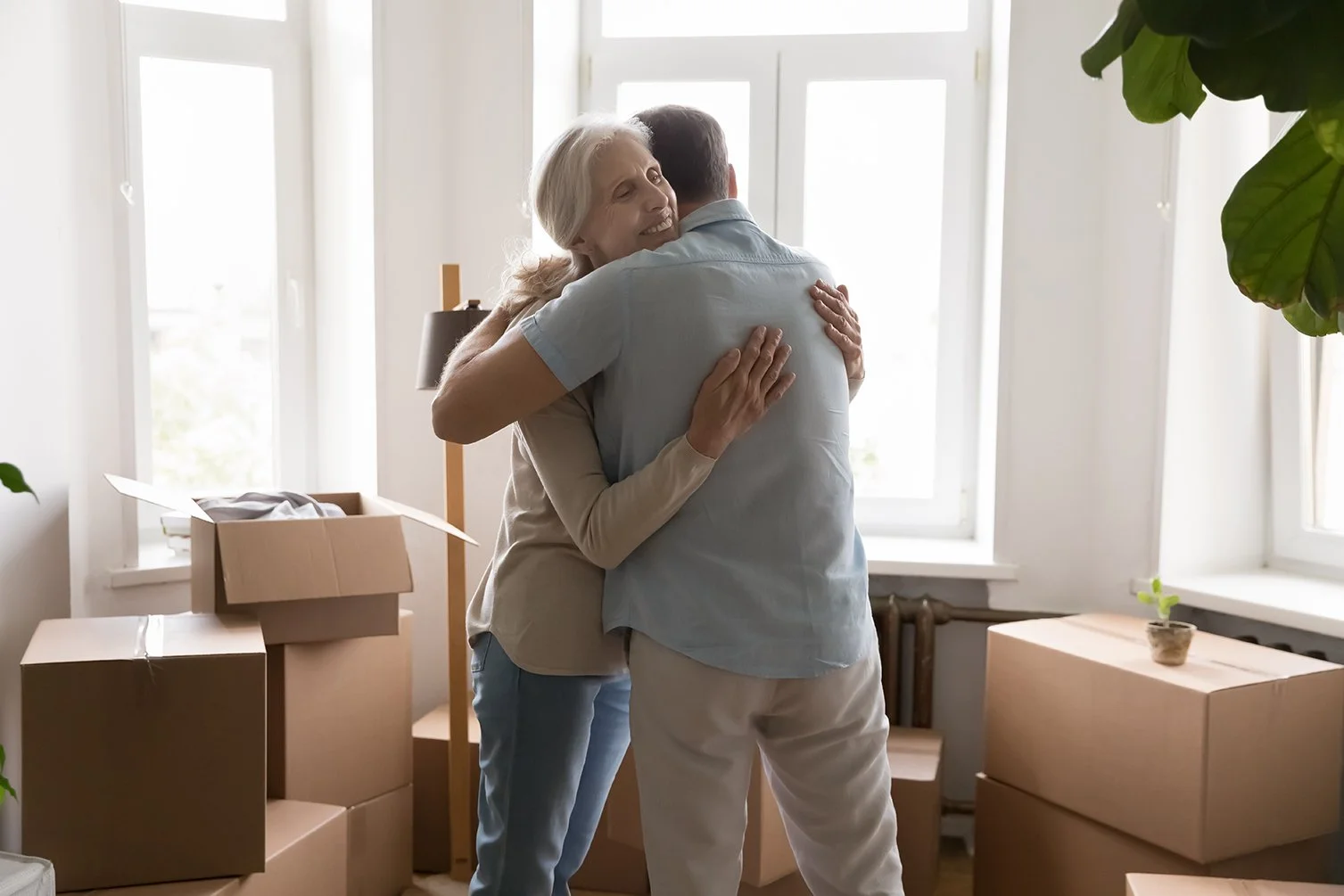 Senior couple hugging in a bright room surrounded by moving boxes during a downsizing move.