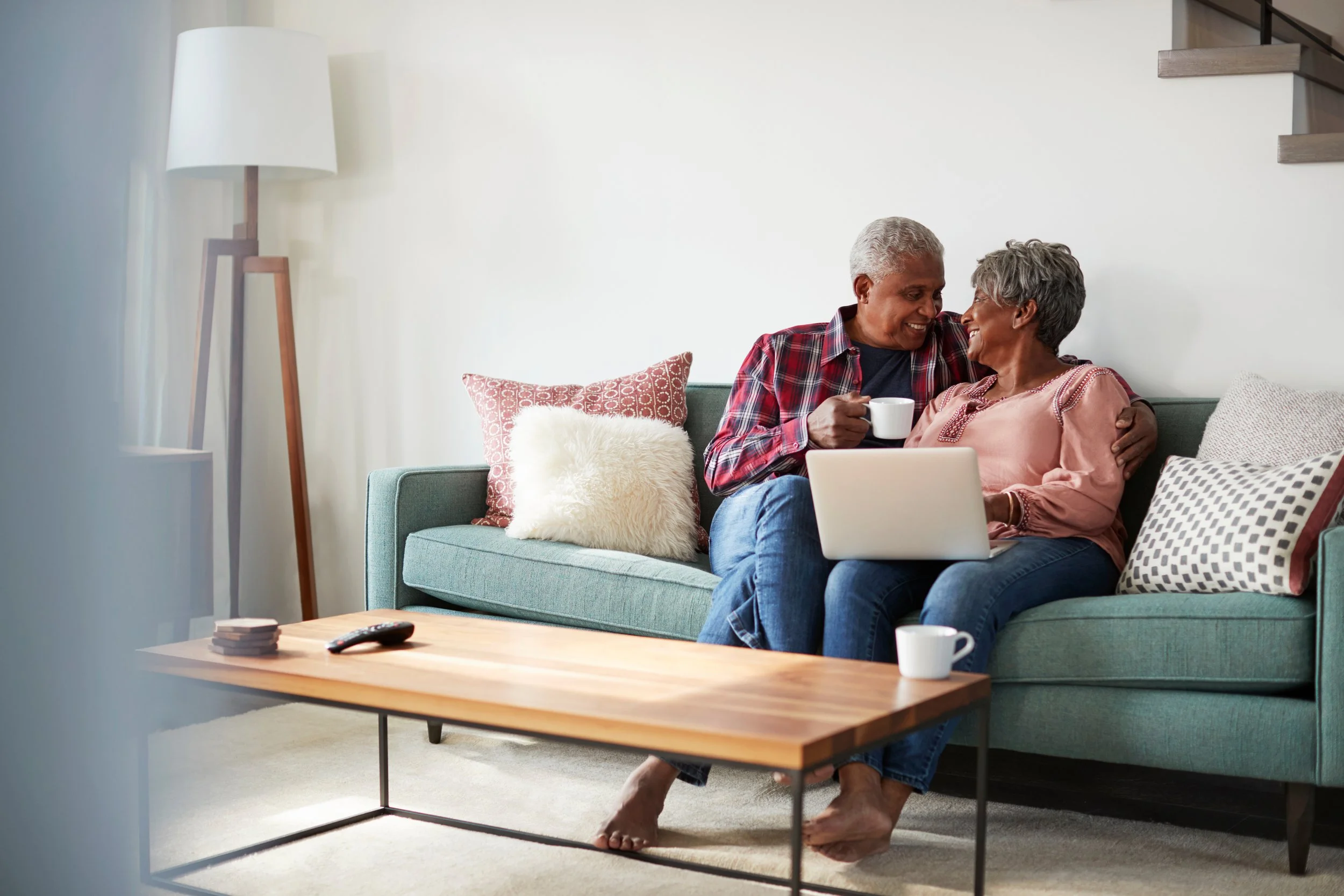 Older couple sitting together on a living room sofa, smiling and looking at a laptop while planning at home.
