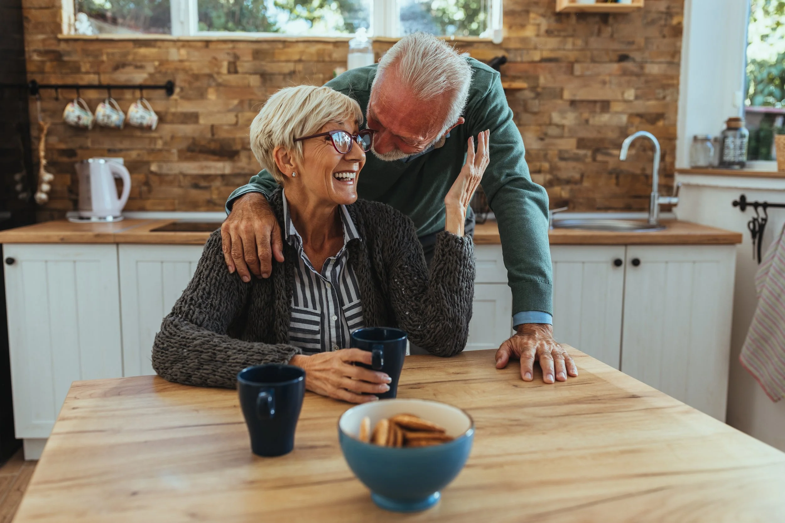 Senior couple sitting at a kitchen table with coffee cups and a bowl of cookies, with one person standing behind the other with an arm around the shoulder in a bright, cozy home kitchen.
