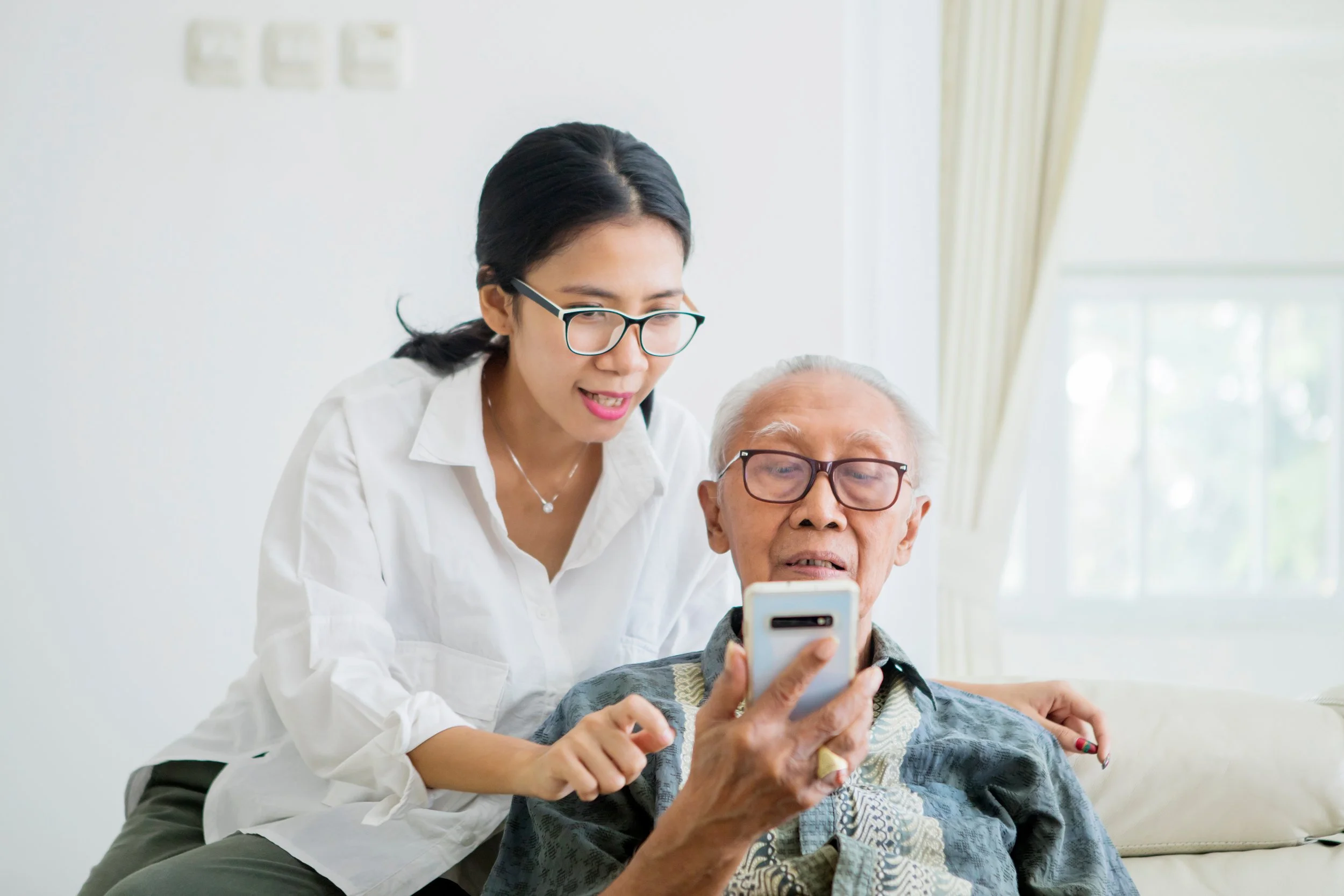 A person sitting on a couch holding a smartphone while another person stands beside them, offering guidance in a bright living room setting.
